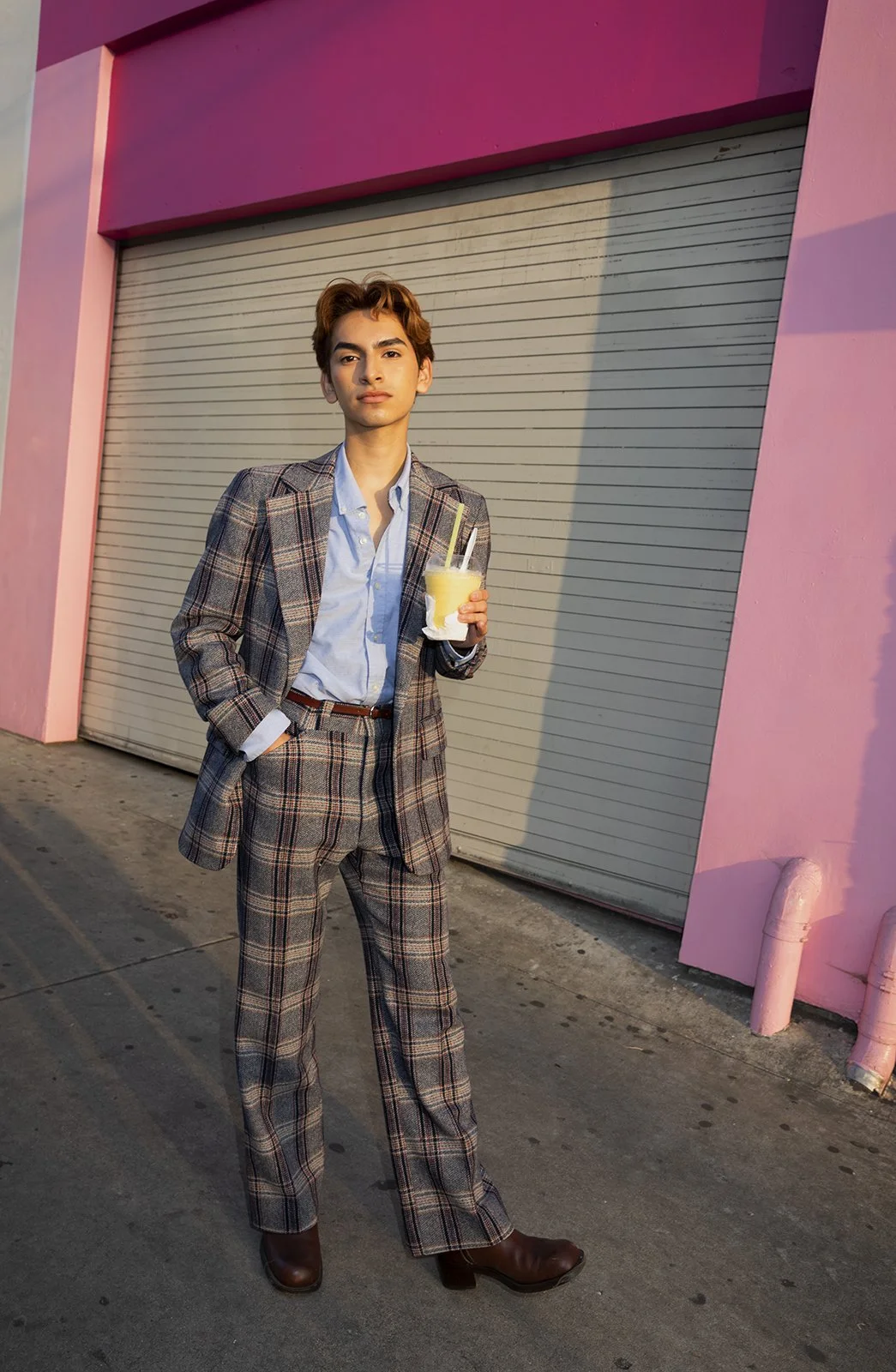 A young man in a plaid suit holds a drink with two straws, standing on a sidewalk in front of a pink building with a closed beige roll-up door.