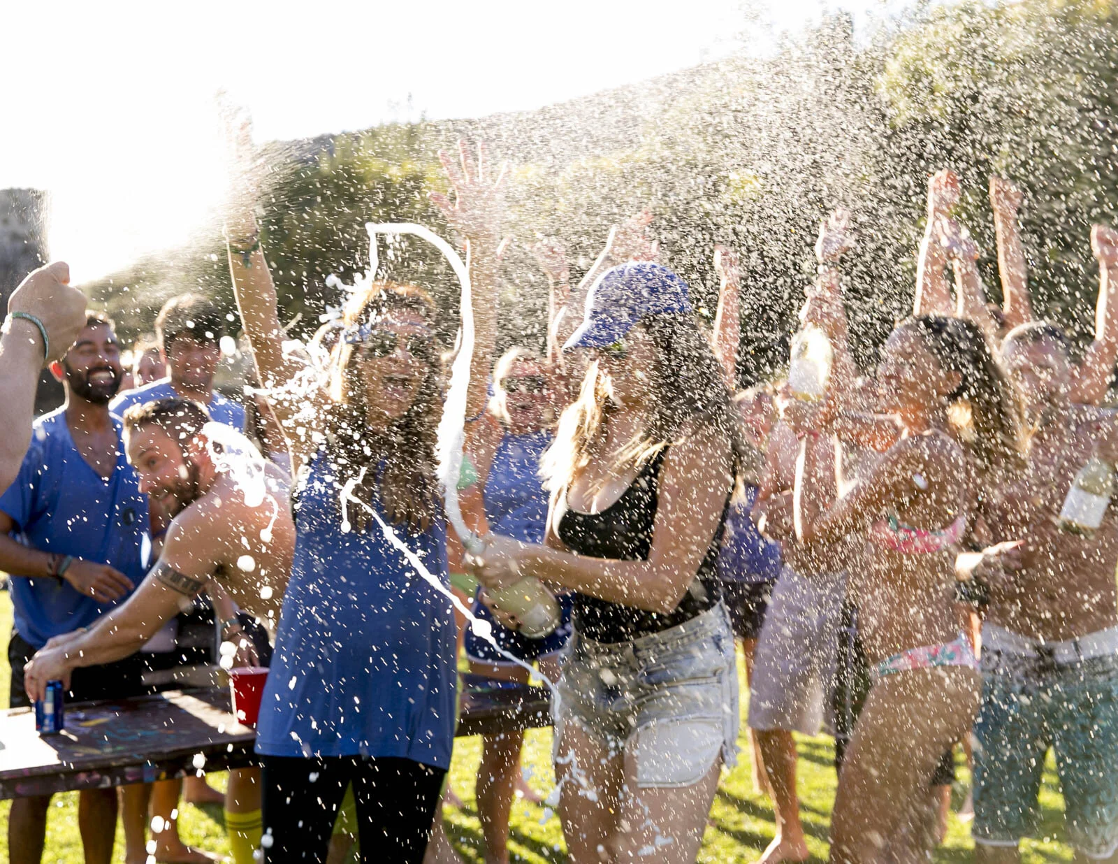 Group of young people having fun at an outdoor party, pouring beer over each other with smiles and raised hands, during daytime.
