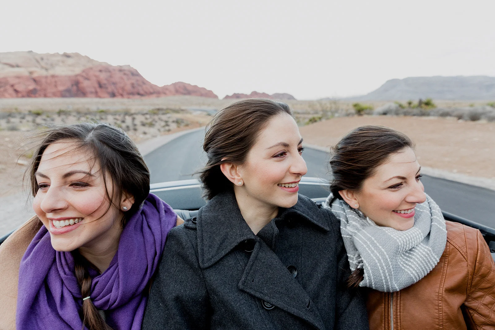 Three women sitting in a car on a desert road, smiling and enjoying the view with red rock formations in the background.