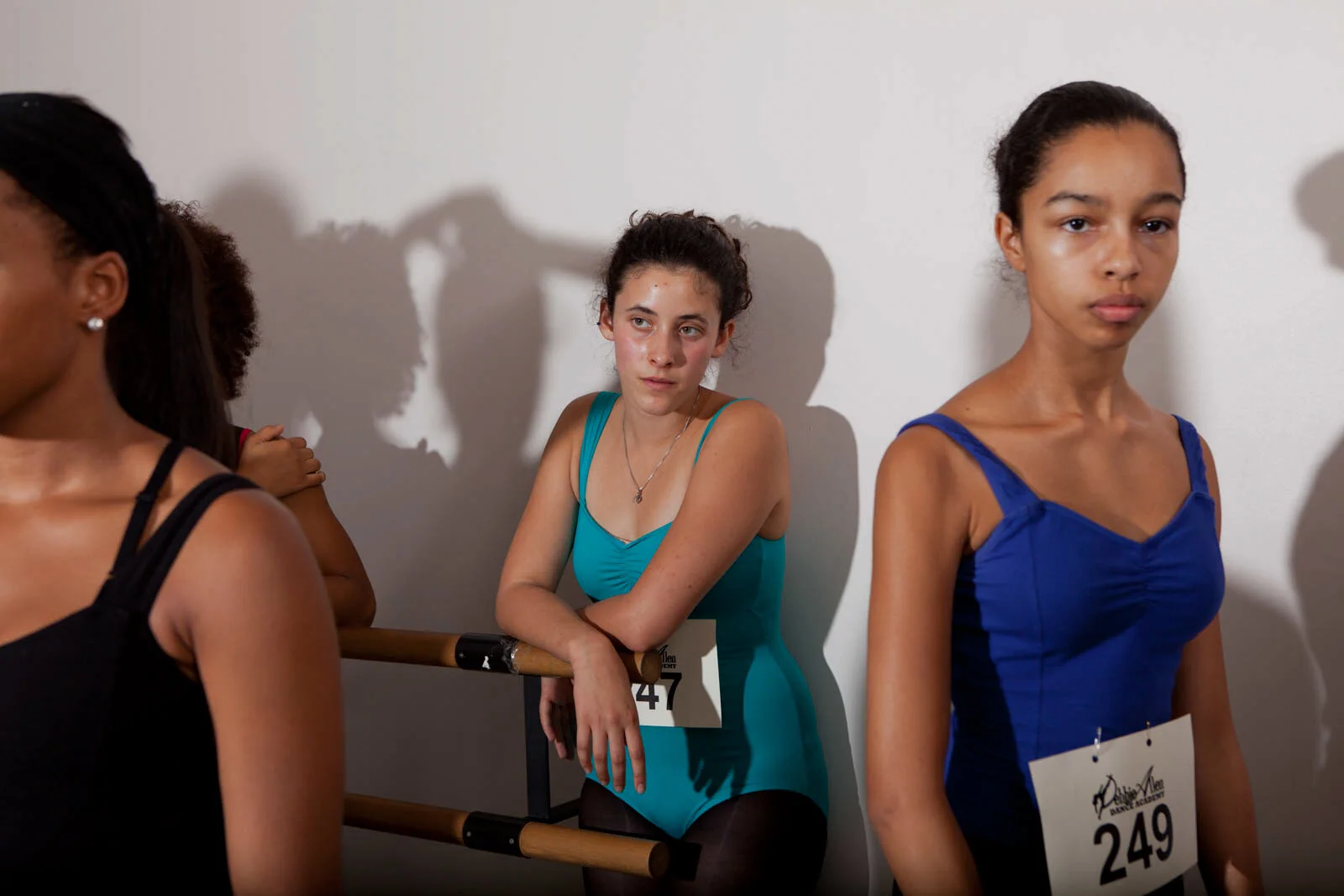 Young women in dance competition waiting behind a ballet bar, some with arms crossed or holding onto the bar, wearing dance attire with contestant numbers displayed, against a plain wall with shadows cast on it.