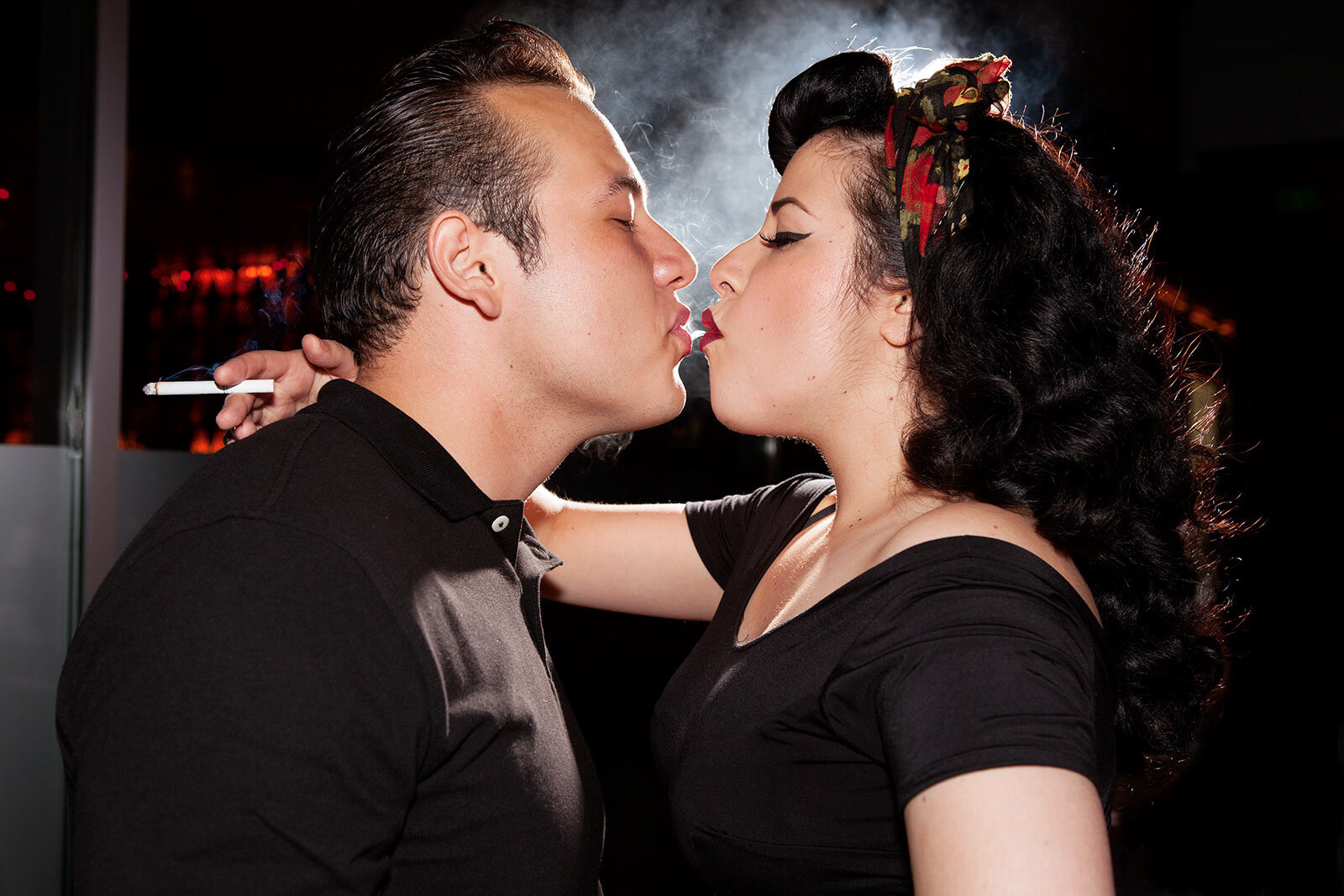 A man and woman are about to kiss, with their eyes closed, in a dimly lit room. The man is holding a cigarette in his right hand.