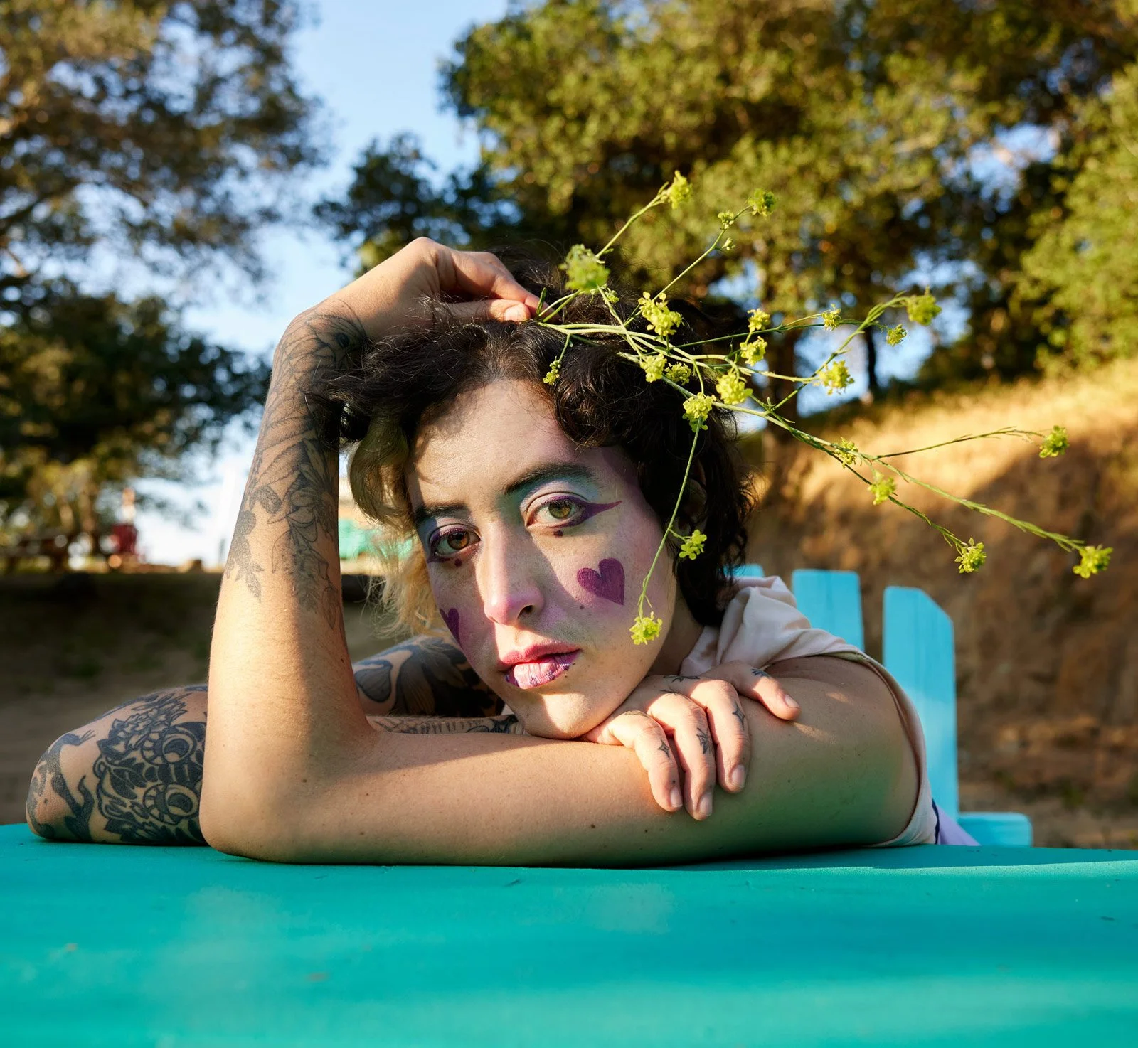 Woman with tattoos and colorful makeup resting her head on her arm outdoors, with yellow flowers on her head and a background of trees and blue sky.