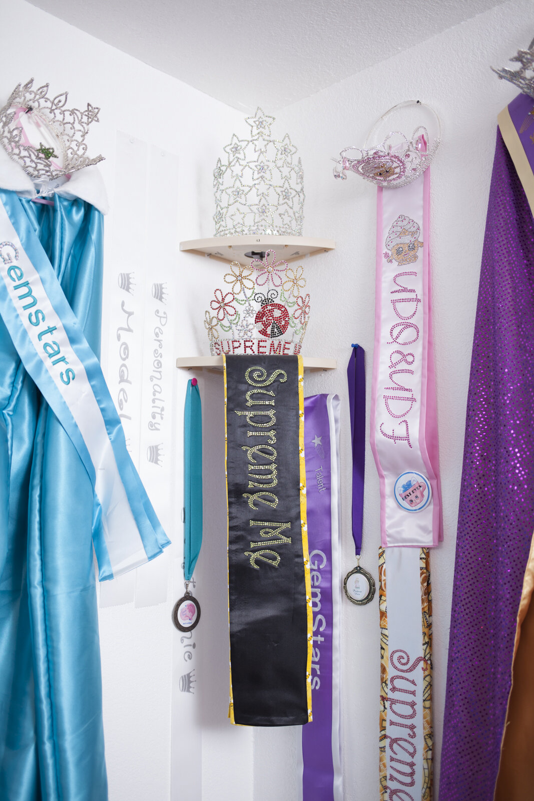 Display of various princess sashes, crowns, medals, and tiaras on a wall, including items labeled 'Princess,' 'Supreme,' and other titles