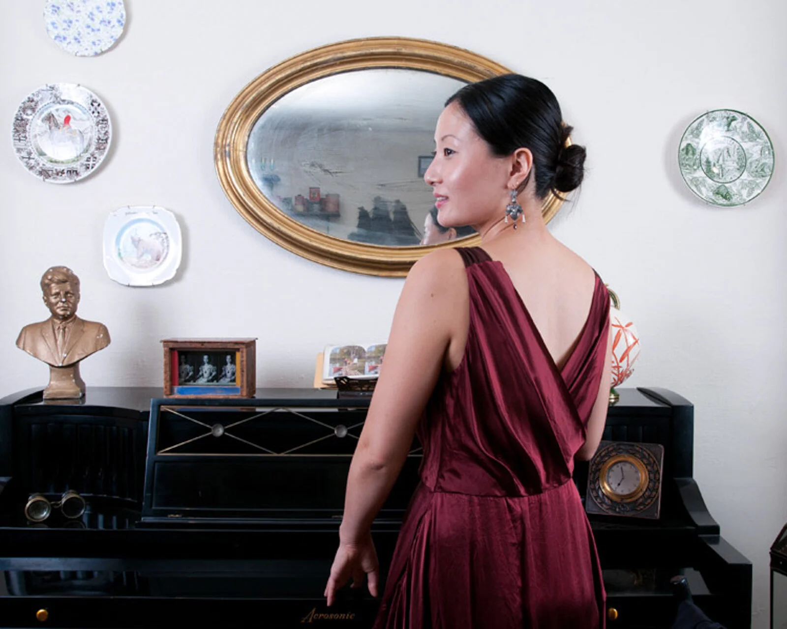 A woman in a red dress standing in front of a piano in a living room, with wall decorations including plates, a mirror, and a bust sculpture.