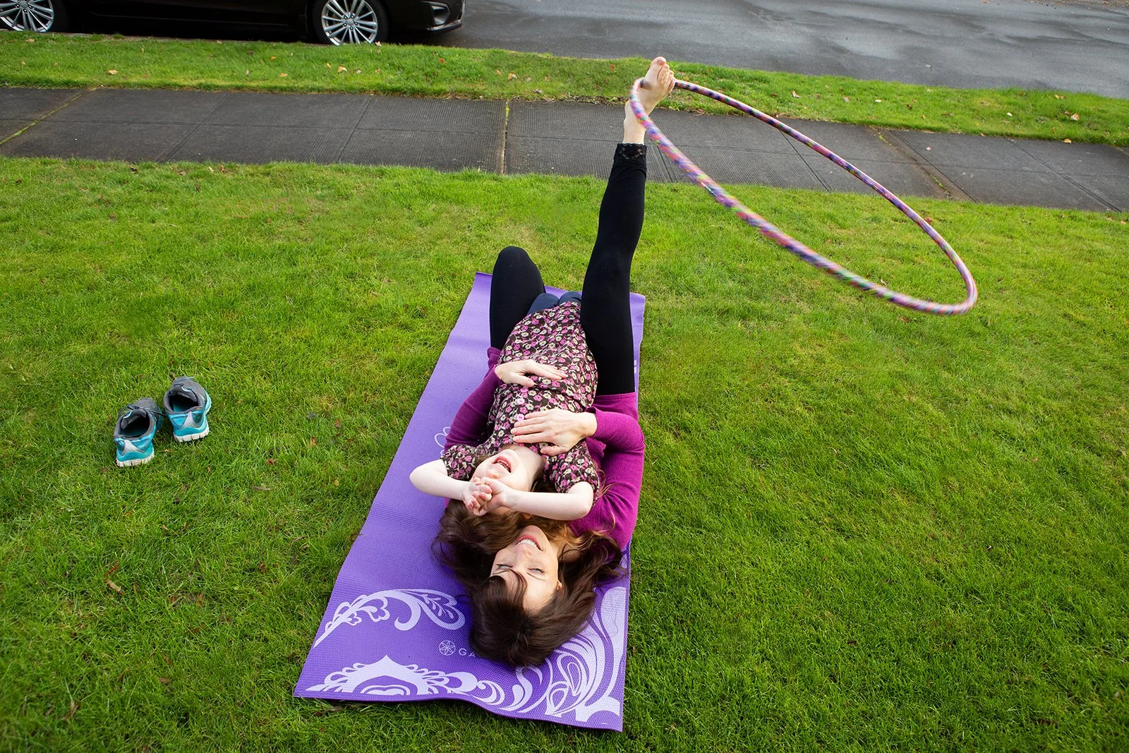 A woman and a young girl lying on a purple yoga mat on a grassy lawn, with the woman holding a hula hoop; shoes are off to the side.