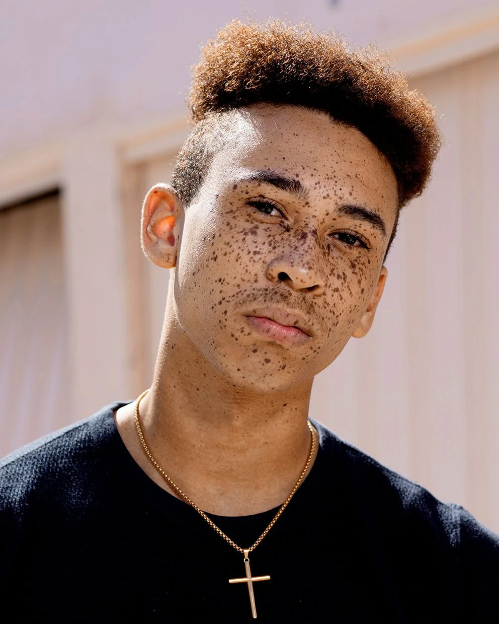Portrait of a young man with curly hair and a prominent amount of freckles, wearing a black shirt and a gold cross necklace, looking at the camera.
