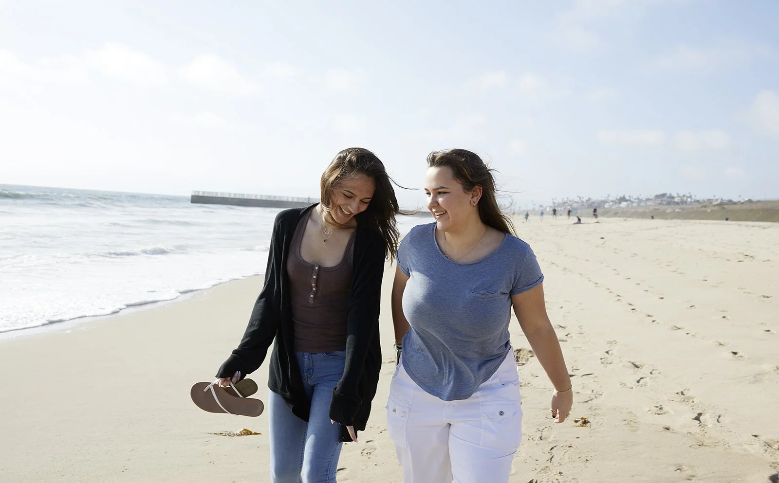 Two young women walking together on a sandy beach, smiling and enjoying the sunny day near the shoreline.