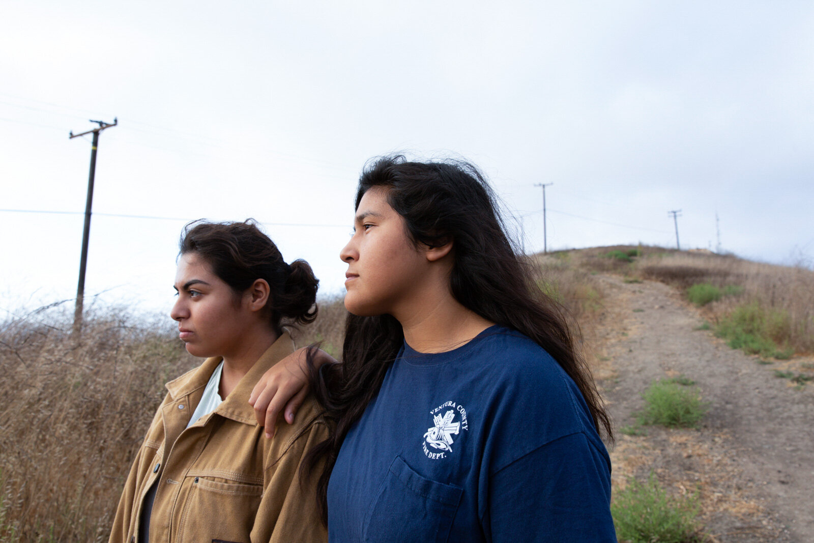 Two women standing side by side outdoors, one with her arm on the other's shoulder, looking serious, with a dirt hill and power lines in the background.