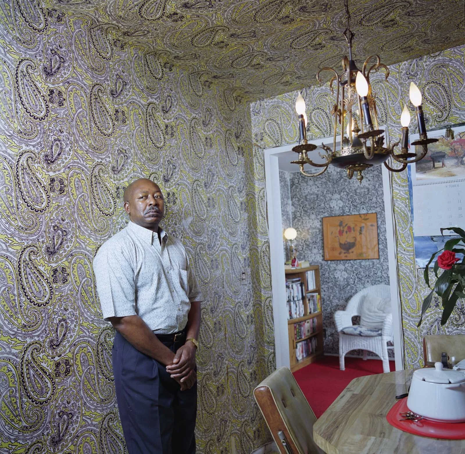 Man standing in a room with vintage paisley wallpaper, next to a doorway leading to another room with floral wallpaper and a white wicker chair.