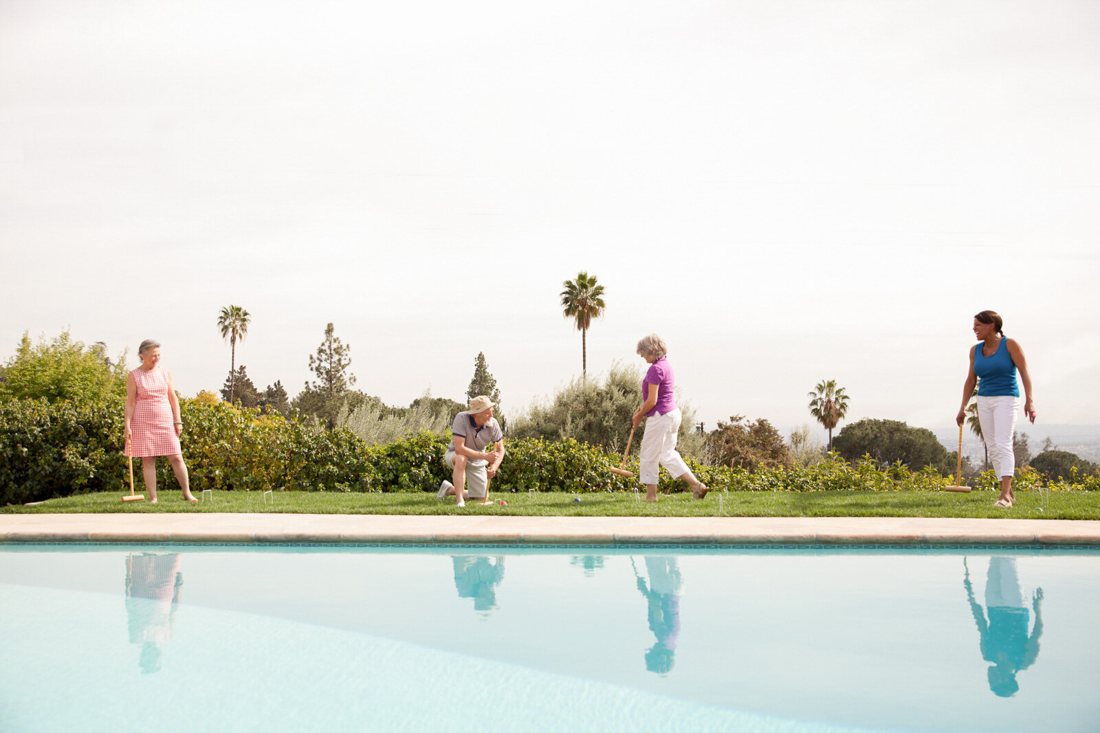 Five women playing croquet by a swimming pool on a sunny day, with trees and palm trees in the background.
