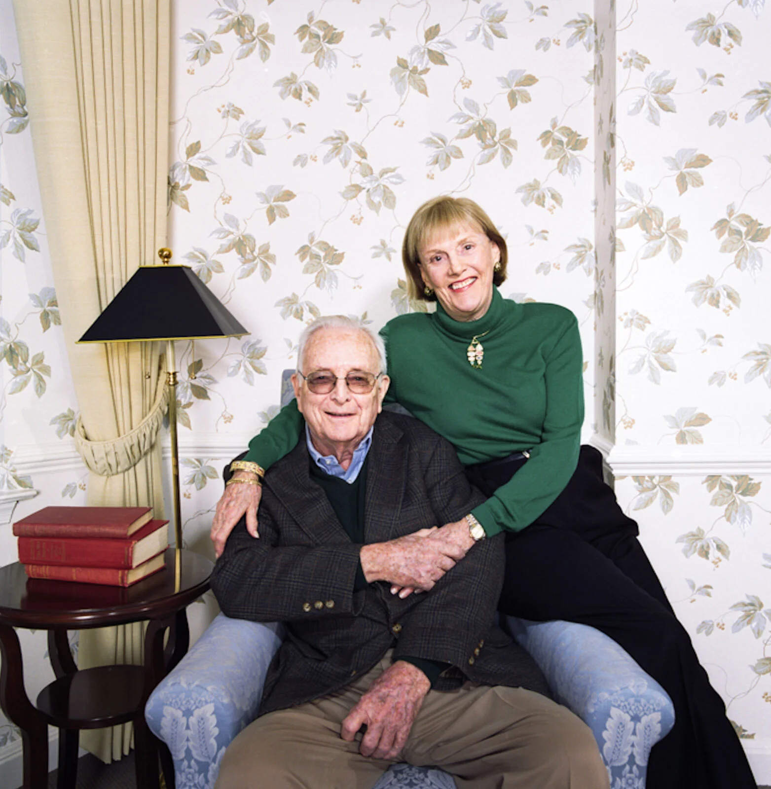 An elderly man and a middle-aged woman, both smiling, posing together in a living room. The man is sitting on a blue armchair, wearing sunglasses, a checkered blazer, and beige pants. The woman is standing behind him with her arm around his shoulders