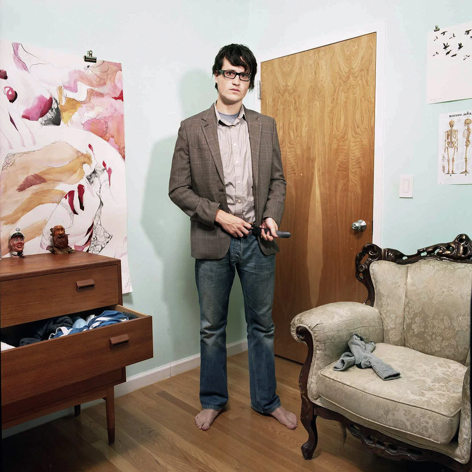 A man with dark hair, glasses, wearing a brown plaid blazer and jeans, standing barefoot in a room, holding a pipe wrench. The room has a vintage armchair with a sock on it, a wooden dresser with dolls, and colorful artwork on the wall.