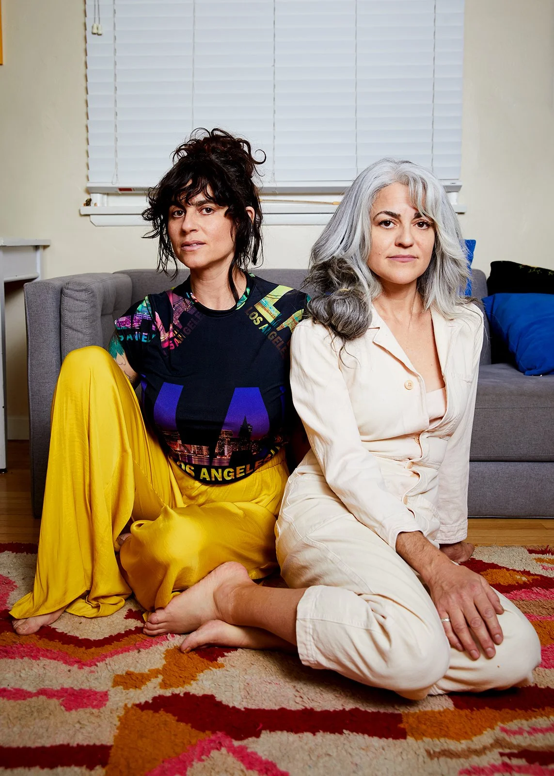 Two women sitting on the floor in a living room, one with dark, curly hair wearing a colorful Los Angeles-themed shirt and yellow pants, and the other with gray hair in a light-colored outfit. They are close to each other, with the woman with gray hair having her hand on the floor.