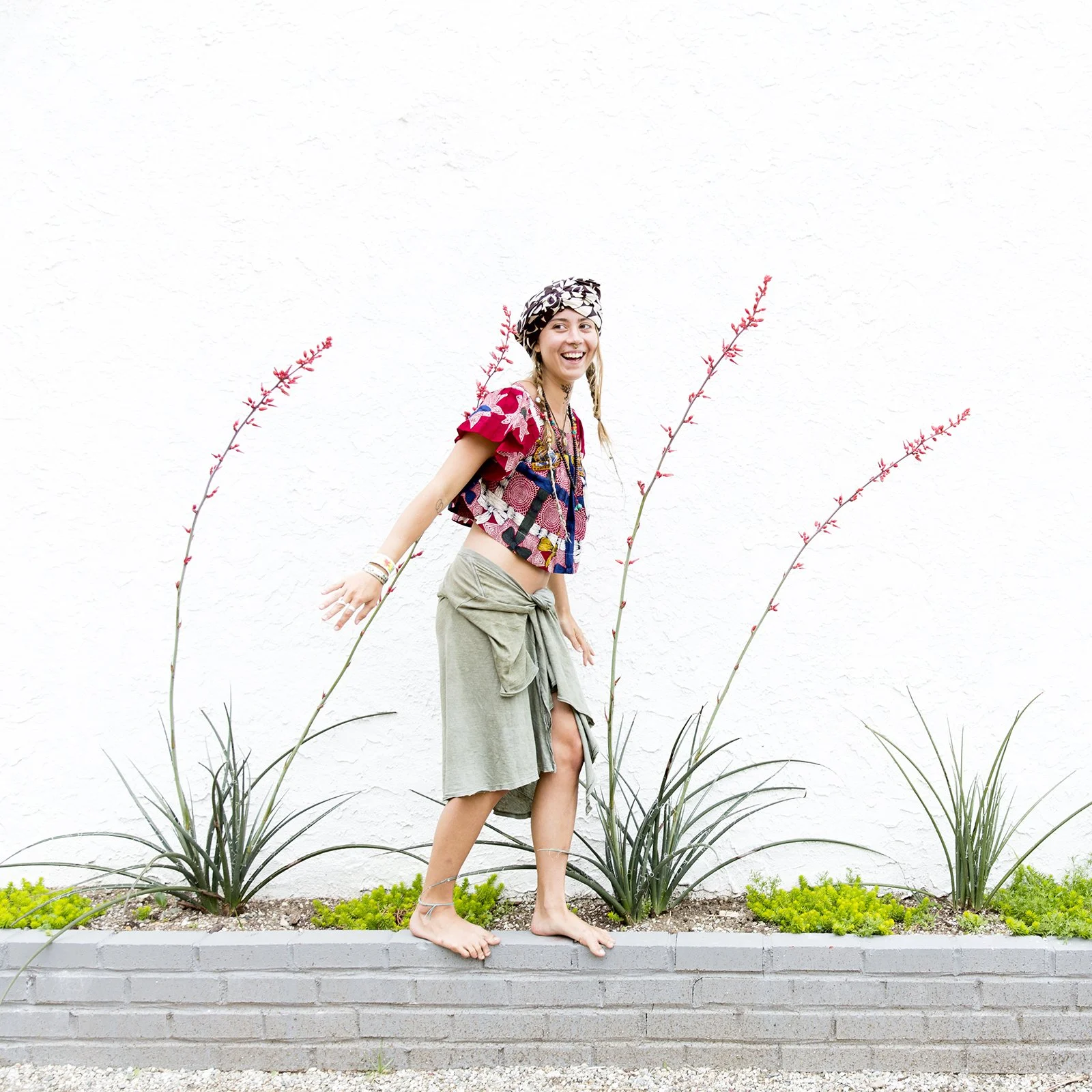 Woman standing barefoot on a brick border next to plants, smiling, wearing a colorful top, a green skirt, a headscarf, and layered jewelry, against a plain white wall.