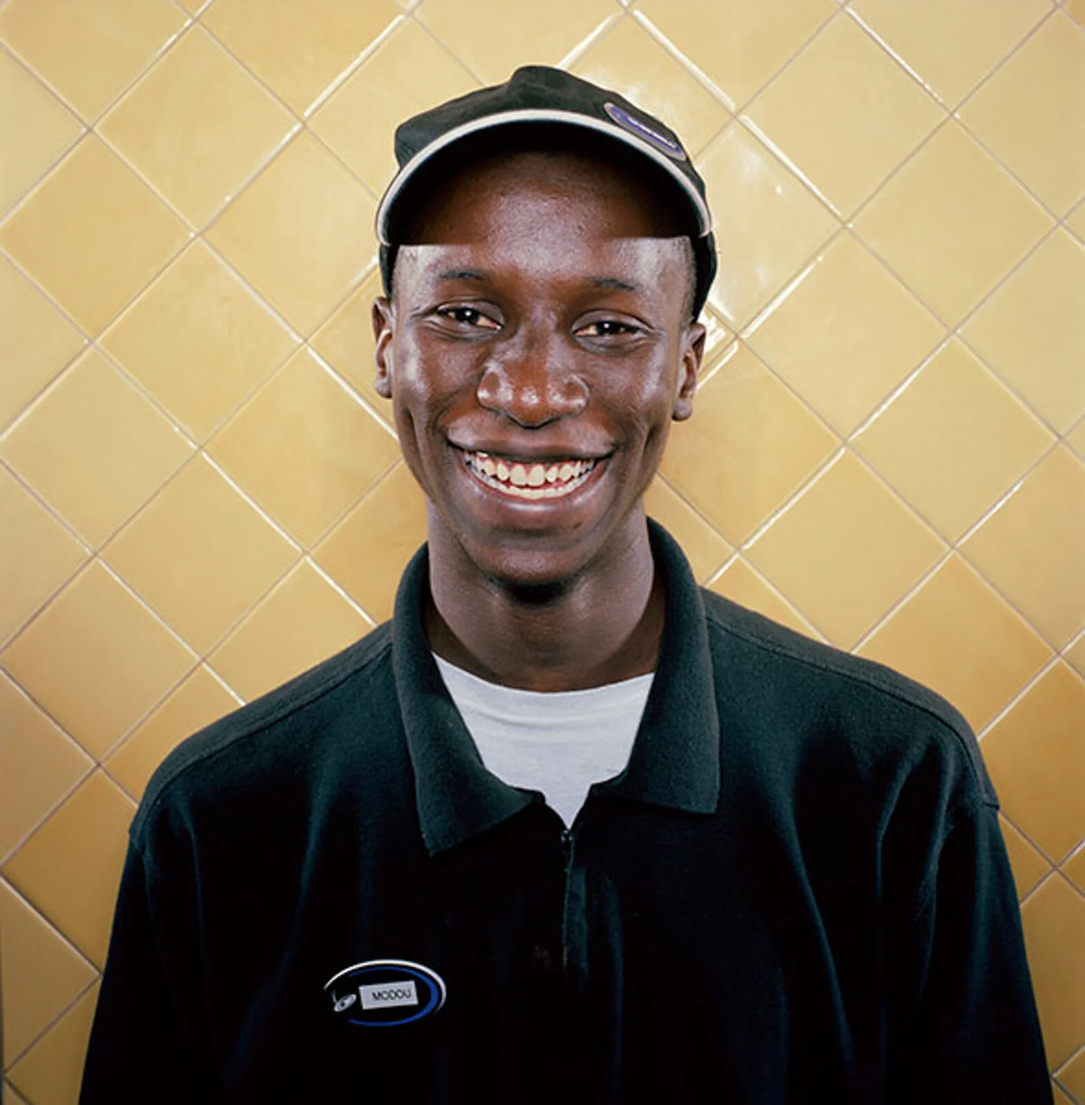 A young man smiling, wearing a black cap, black jacket, and a white shirt with a name tag, standing in front of a yellow tiled wall.