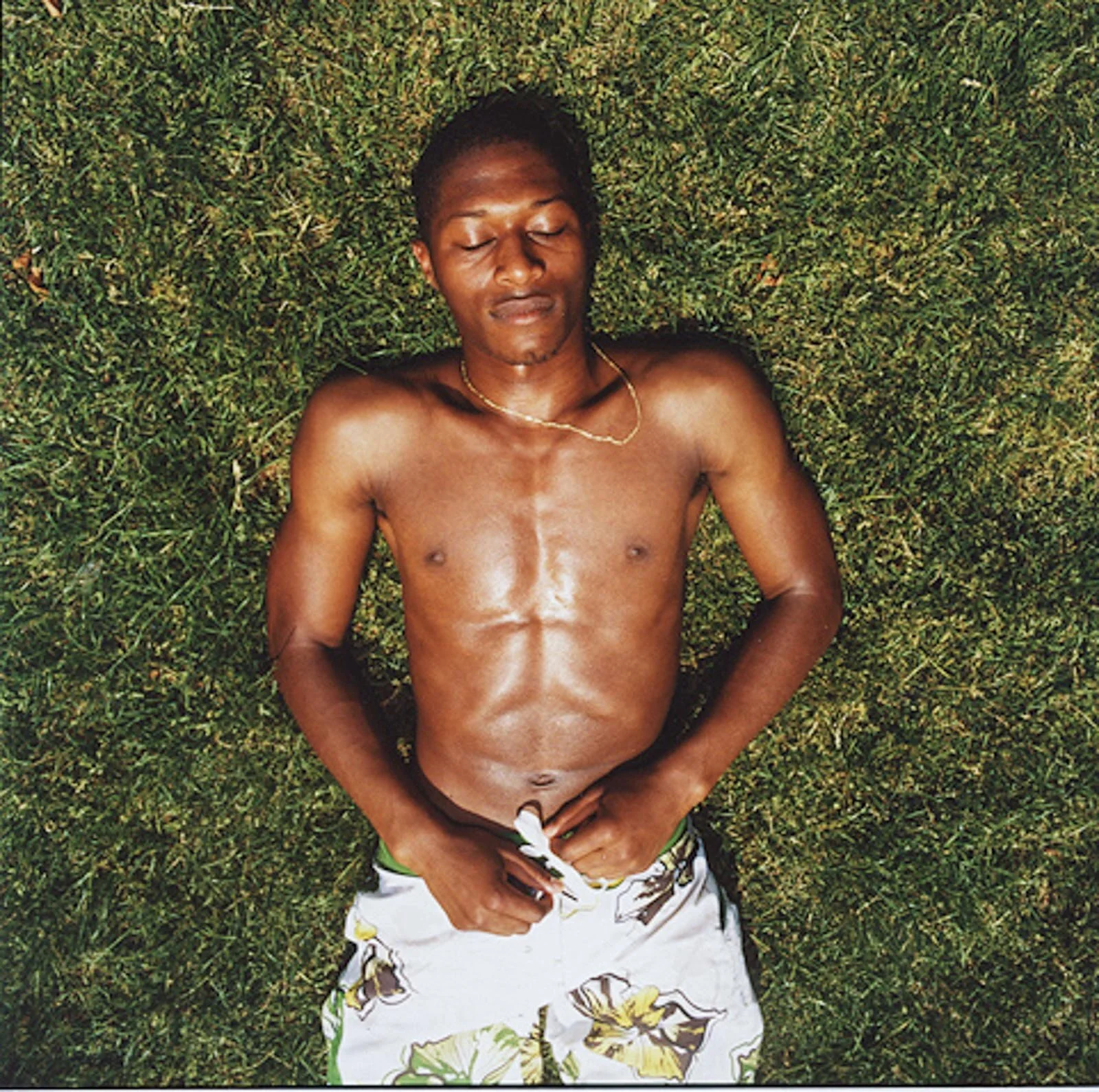 A shirtless young man with dark skin and short black hair lying on green grass with eyes closed, wearing floral-patterned shorts and a gold chain necklace.