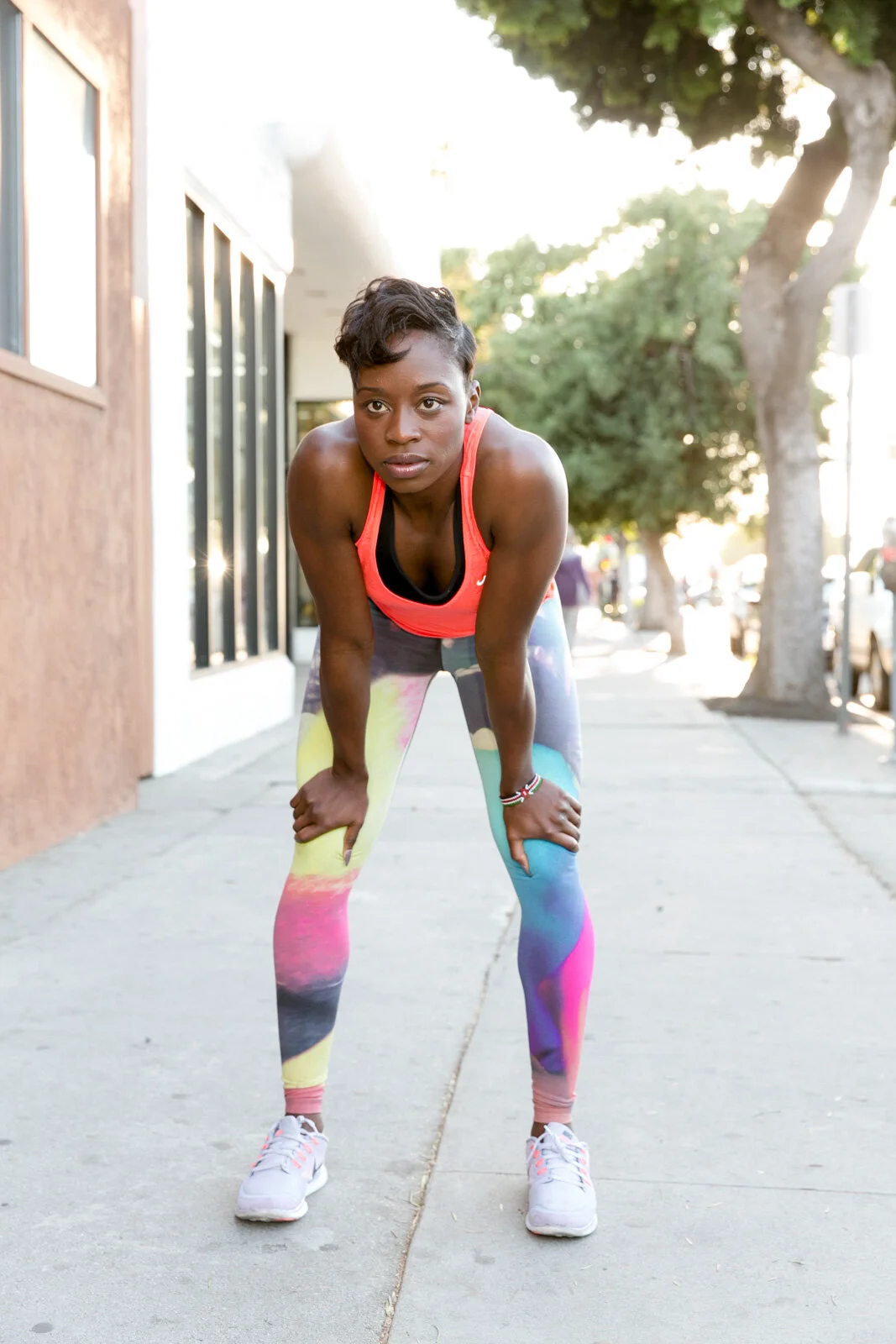 A woman in colorful athletic leggings and a pink tank top leaning forward on a city sidewalk after exercising.
