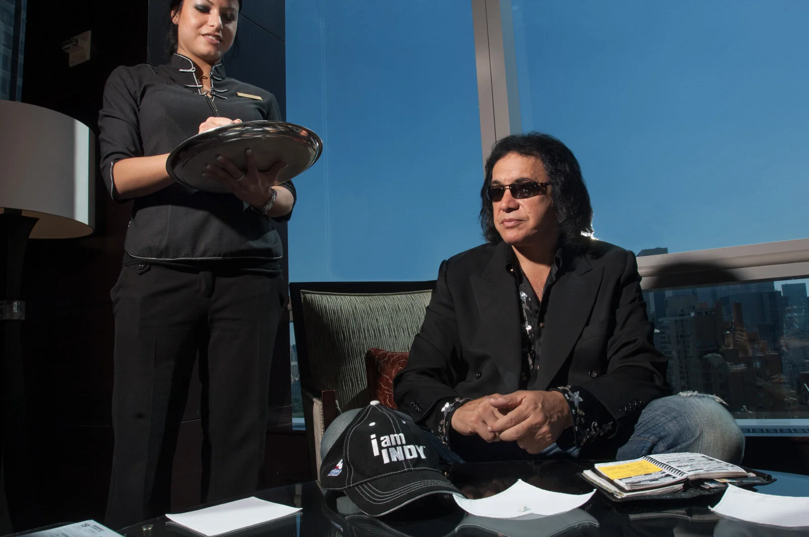 A man with long black hair, wearing sunglasses and a black blazer, sitting at a desk with a cap that says 'I am INDY' on it, and a woman in a black uniform standing nearby, holding a tray, in an office with large windows showing a cityscape in the ba