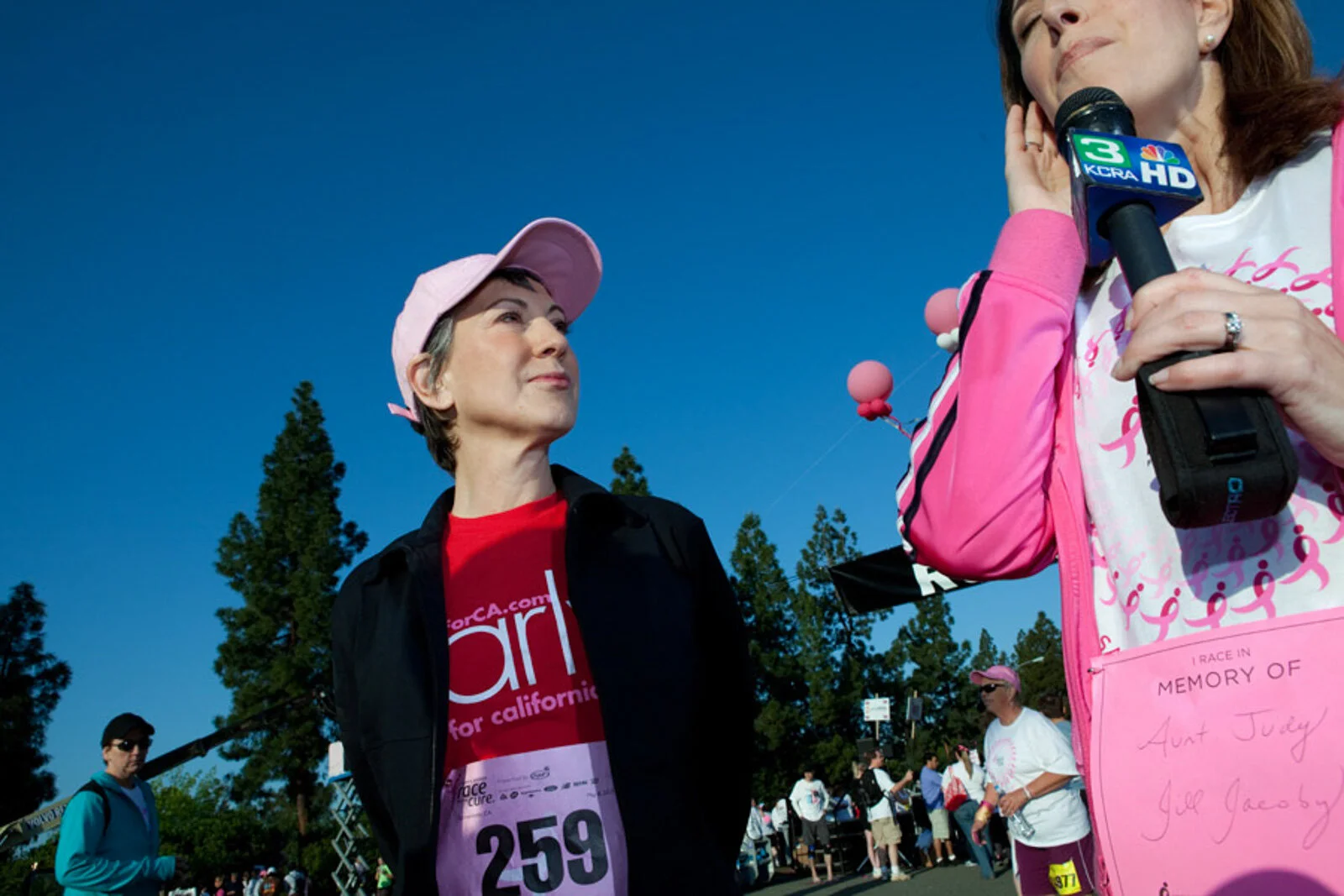 A woman being interviewed at a charity race event, with numerous participants in the background, some wearing pink shirts and hats, and a blue sky overhead.