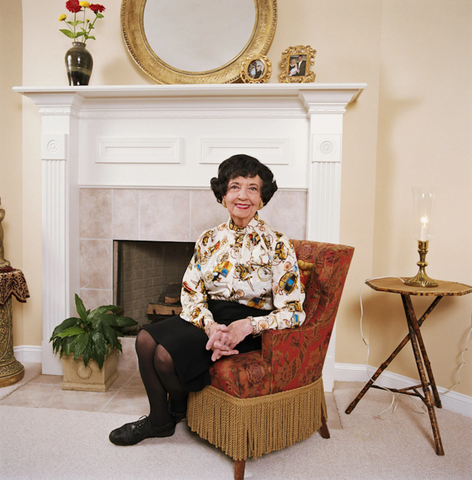 An elderly woman sitting on a patterned red and gold armchair in front of a fireplace, wearing a white blouse with a colorful pattern, black skirt, and black shoes, smiling at the camera. A plant is on the floor to her left, with a table holding a ca