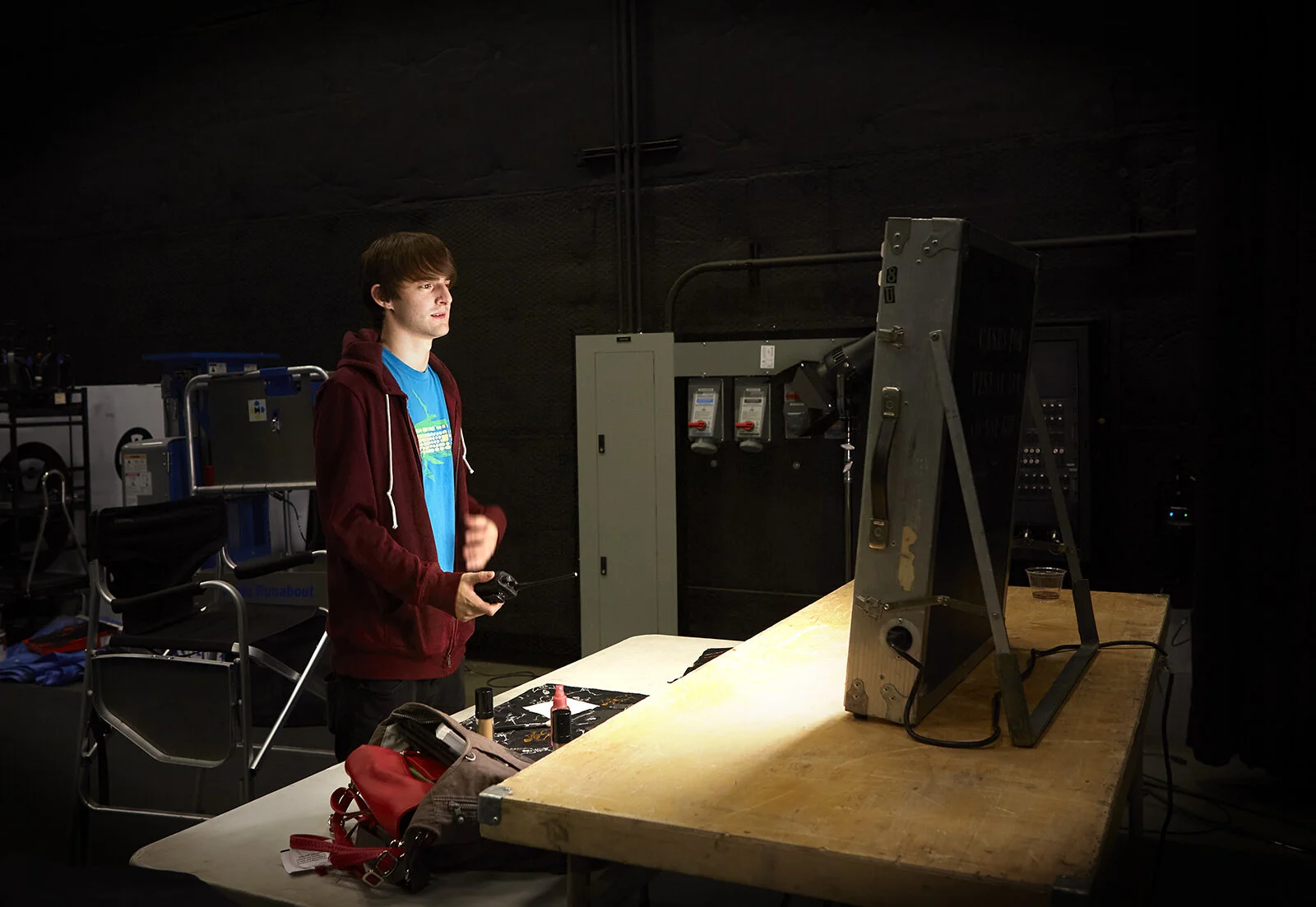 A young man with brown hair wearing a red hoodie and blue t-shirt operating a robot in a workshop.
