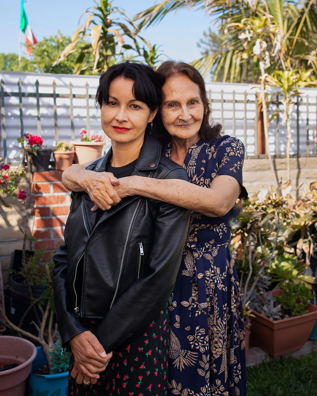 An elderly woman hugs a younger woman in a garden with potted plants, a brick wall, and a white fence in the background.