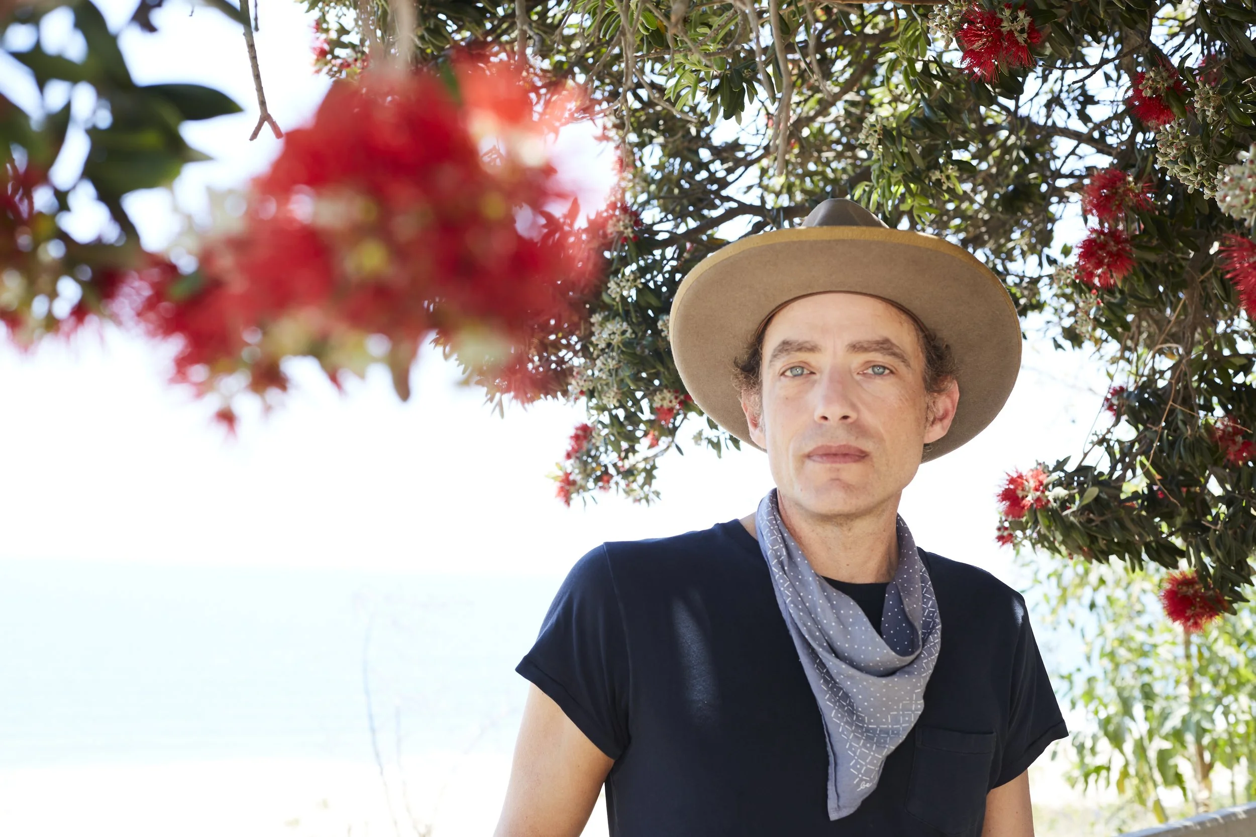 A man wearing a wide-brimmed hat and a dark shirt stands outdoors near flowering red and white blossoms, with the sky and water in the background.