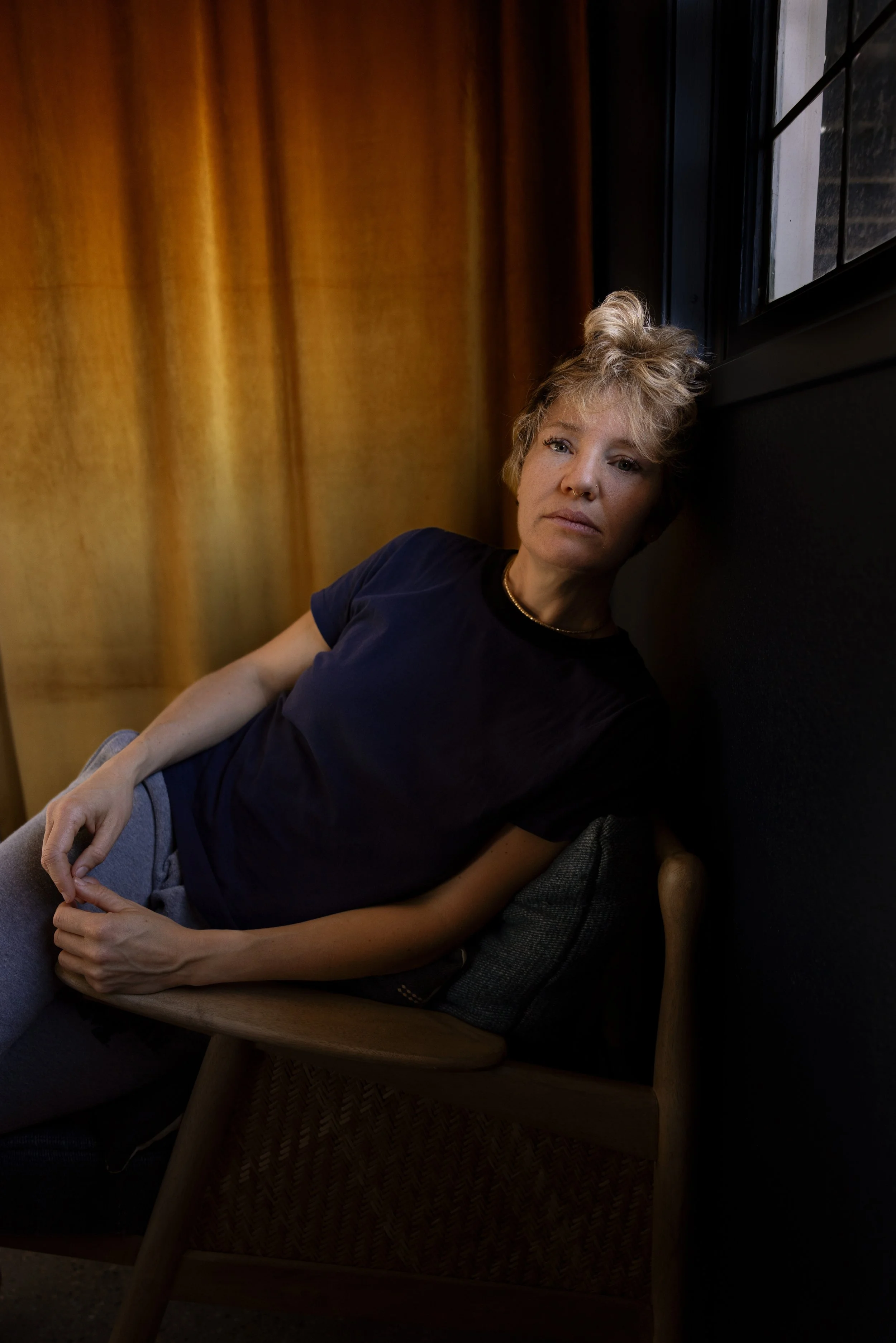 A woman with curly blonde hair resting against a wall, sitting on a wooden chair, near a window with black framing, in a room with wooden wall panels.
