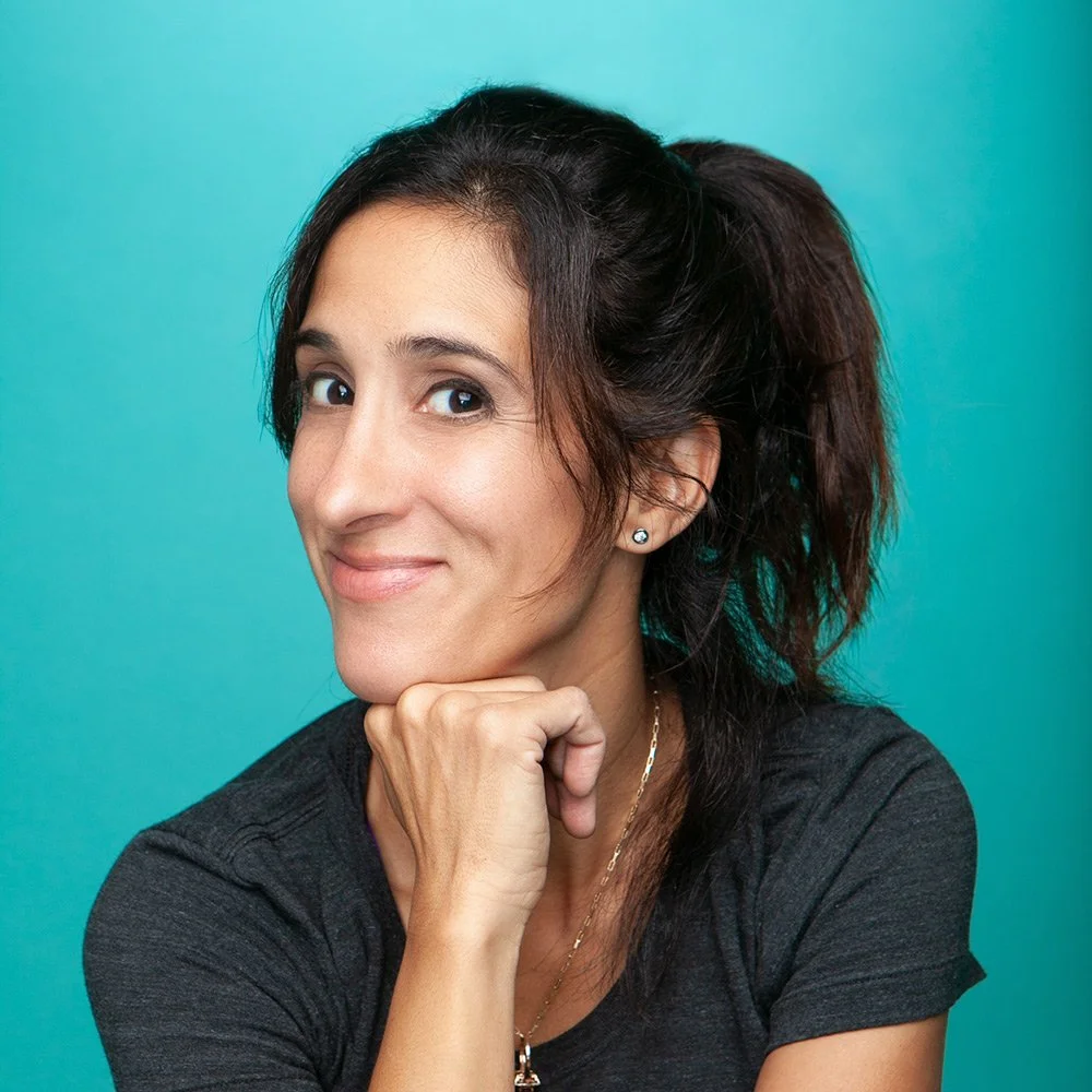 A woman with dark hair tied back, wearing a black t-shirt, earrings, and a necklace, resting her chin on her hand against a teal background.