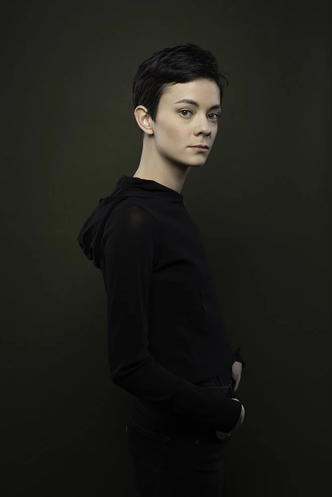 Young woman with short dark hair, wearing a black hoodie and jeans, standing against a plain dark green background, looking at the camera with a neutral expression.