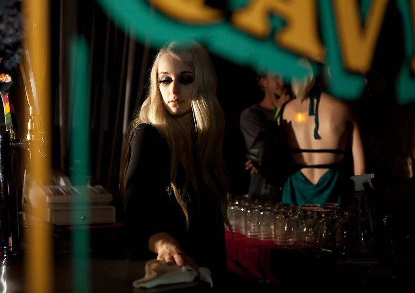 A young woman with long blonde hair and dark makeup standing at a dimly lit bar, with blurred people and glasses in the background.