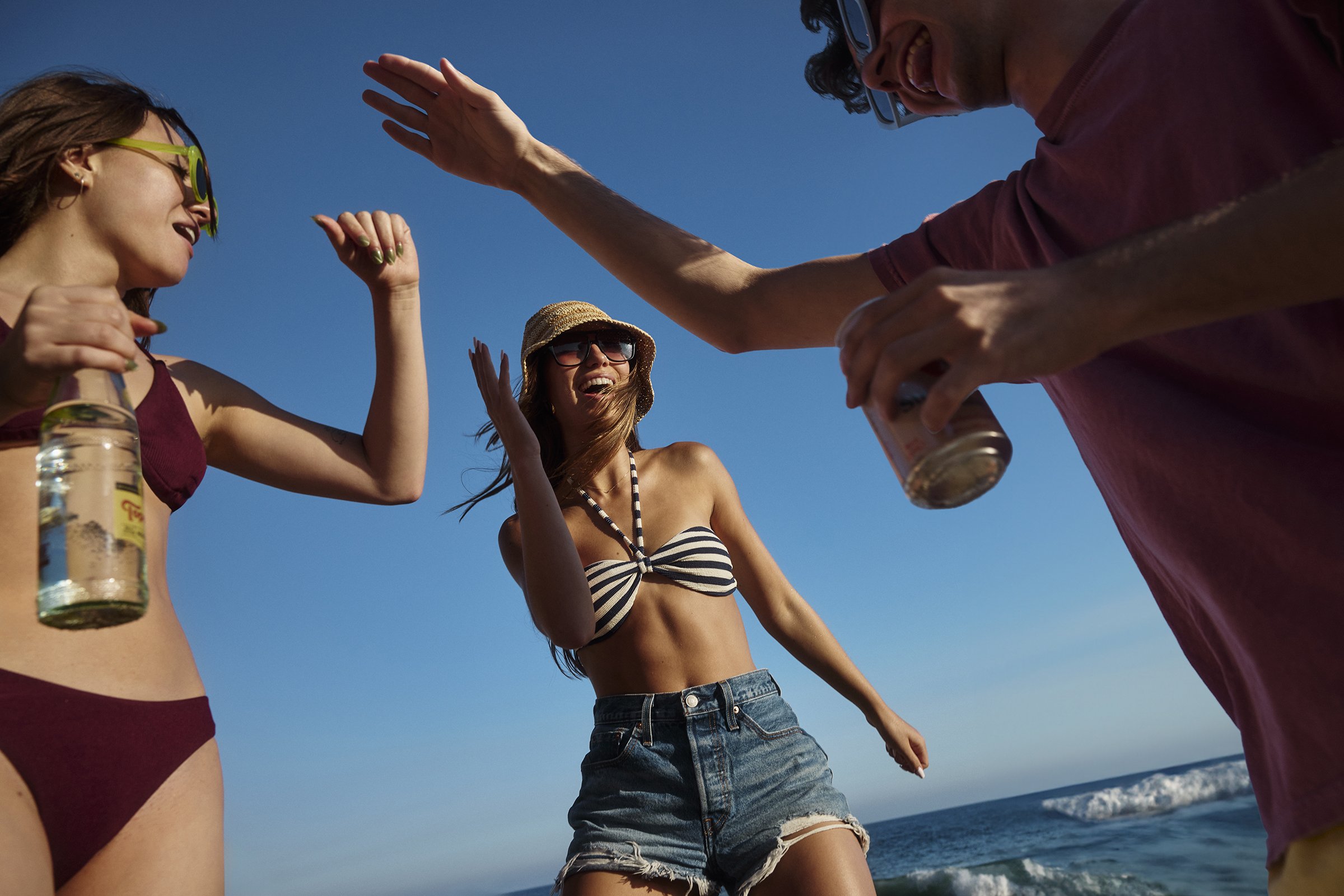 Group of young people enjoying a day at the beach, dancing and smiling under a clear blue sky.
