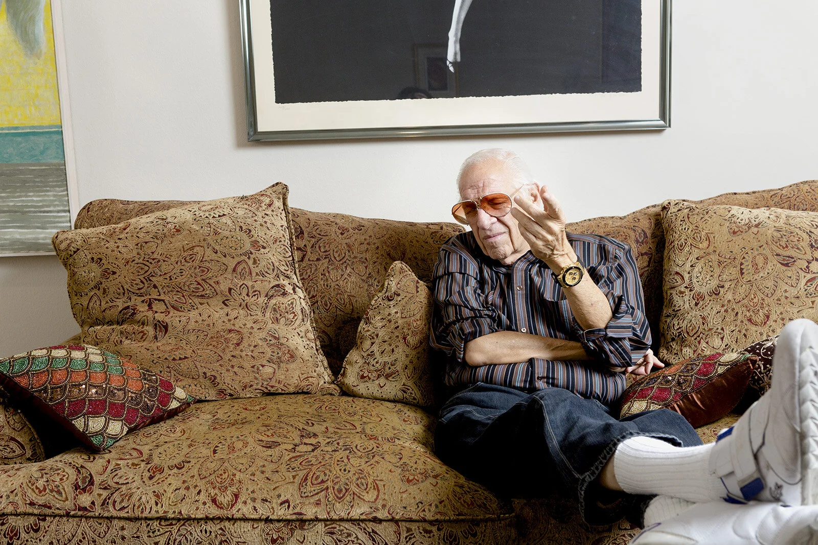 An mature man with white hair wearing sunglasses, a striped shirt, and a watch, sitting on a patterned sofa with multiple cushions, gesturing with his hand. The setting appears to be a living room.