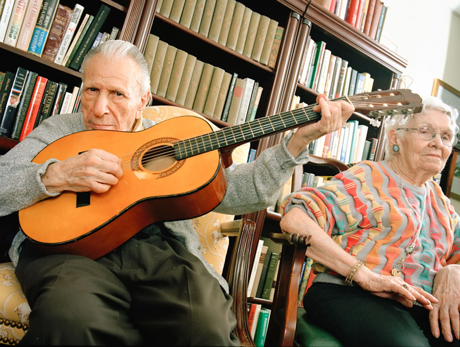 An elderly man playing a guitar while sitting next to an elderly woman in a cozy room filled with bookshelves.