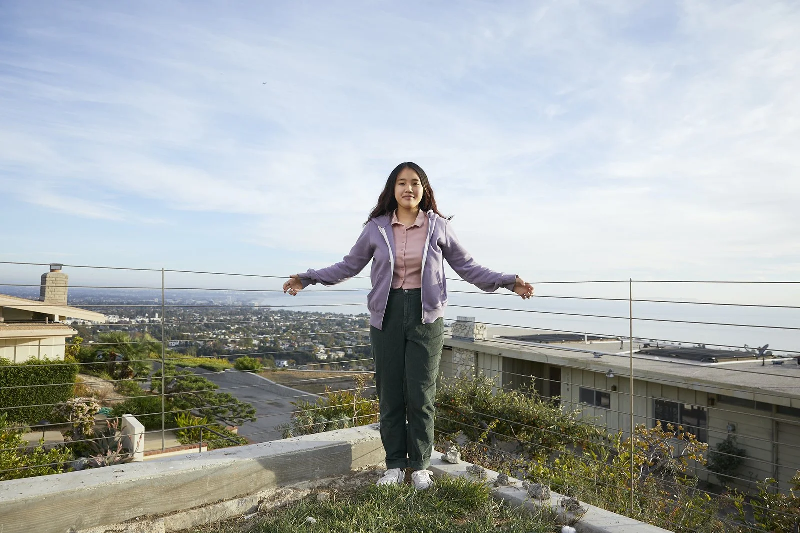 Young woman standing on a rooftop with a city skyline in the background, holding onto metal railing, wearing a purple jacket, pink shirt, green pants, and white sneakers.