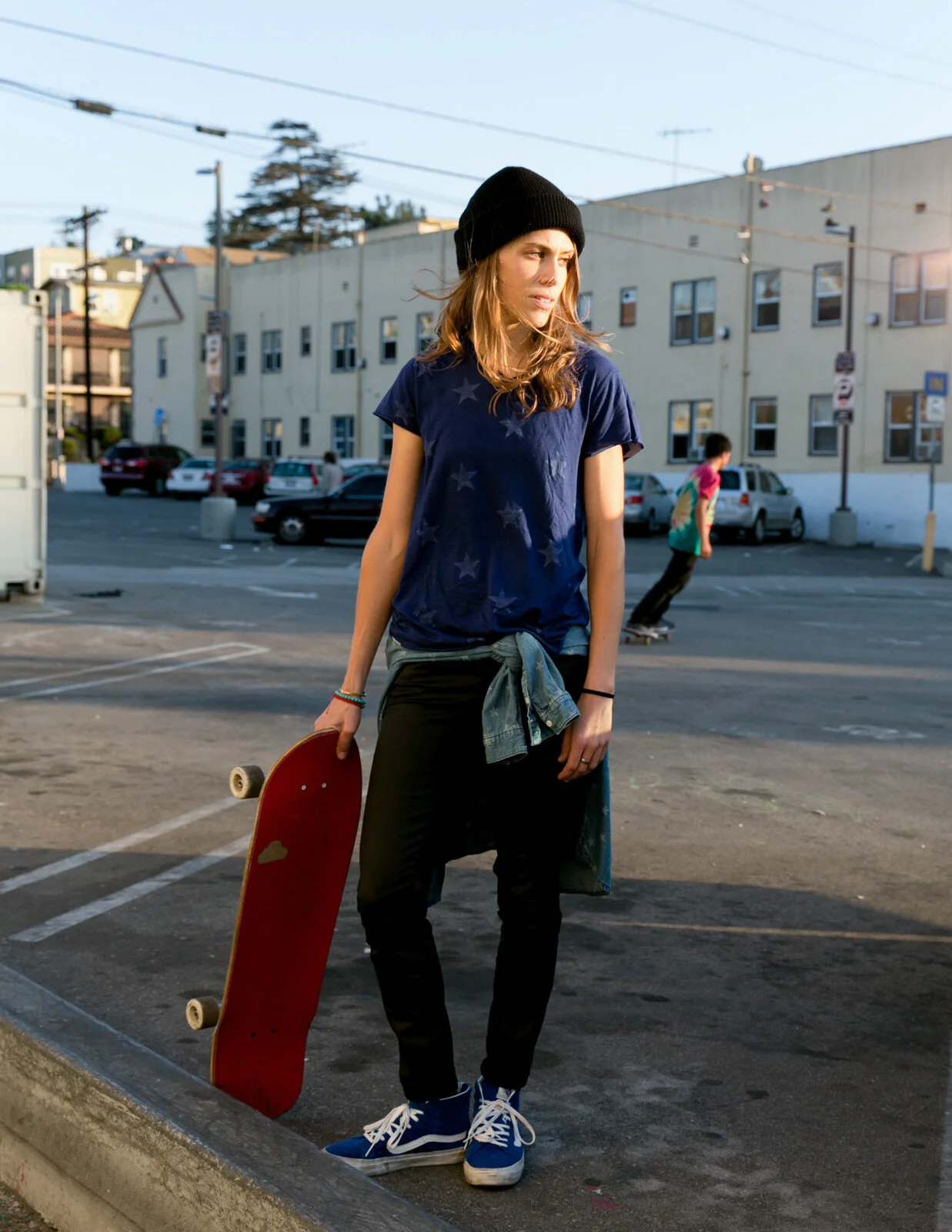 A young woman with a skateboard, wearing a black beanie, navy T-shirt with star print, black pants, and blue sneakers, standing in an urban parking lot with a building in the background.