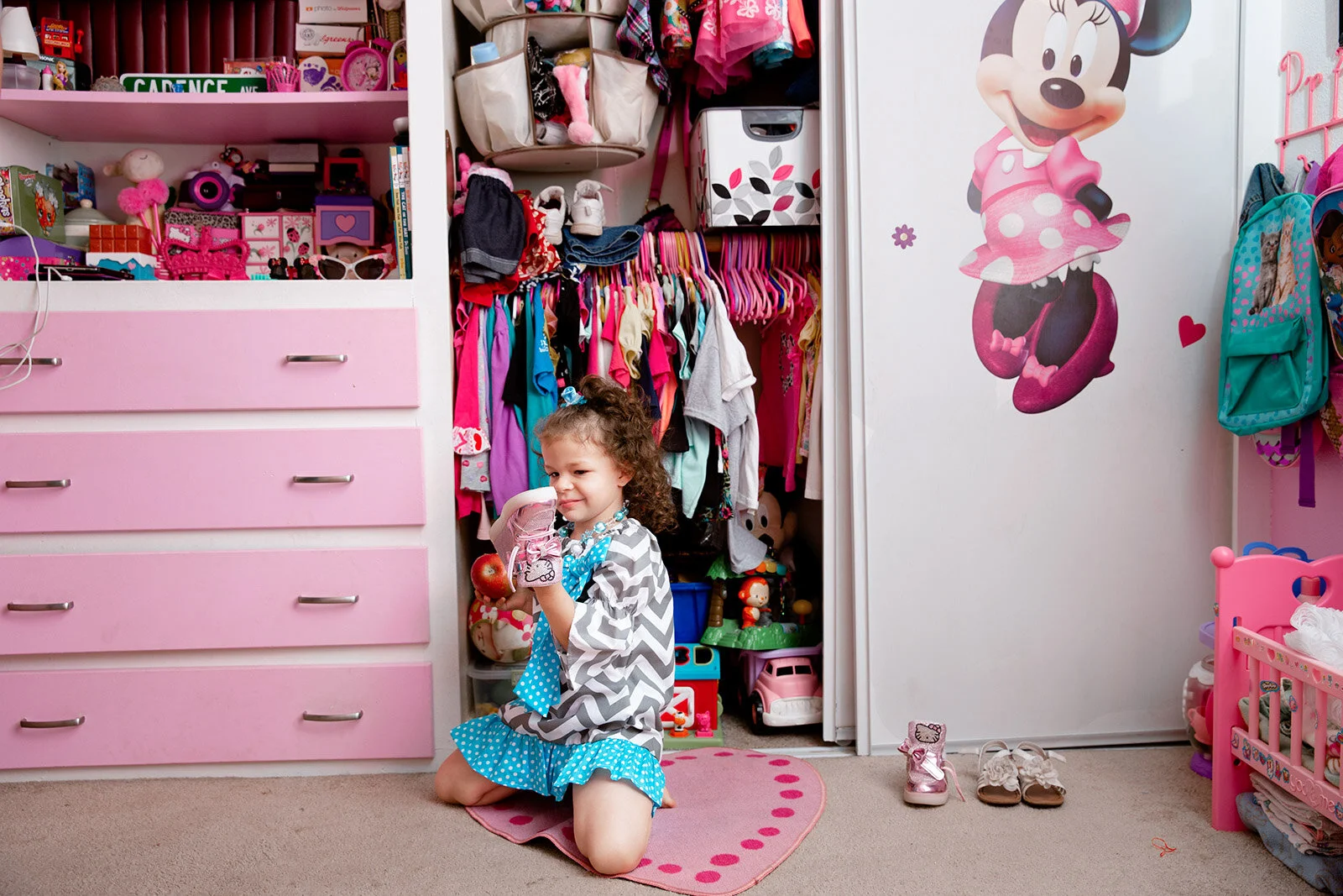 Young girl kneeling on a pink heart-shaped rug in a cluttered children's bedroom, holding a drink and a red apple. The room has pink and white furniture, toys, and clothing, with a large Minnie Mouse sticker on the white closet door.