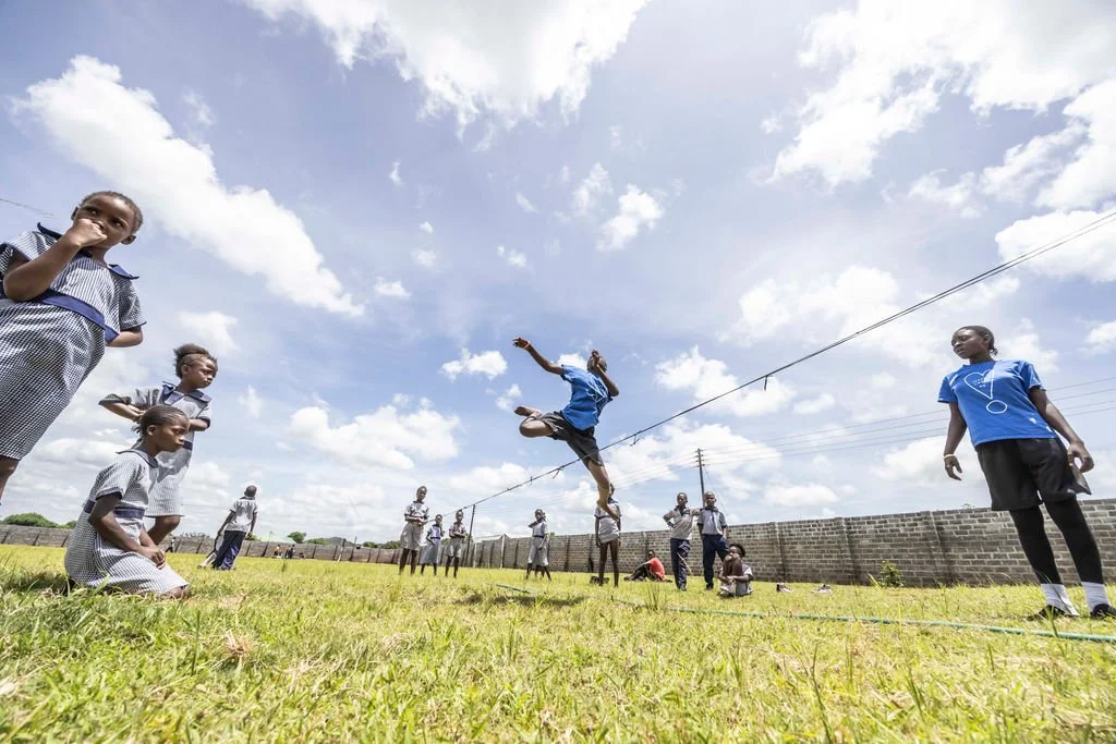 A group of children playing jump rope outdoors on a dry grass field with people and buildings in the background.