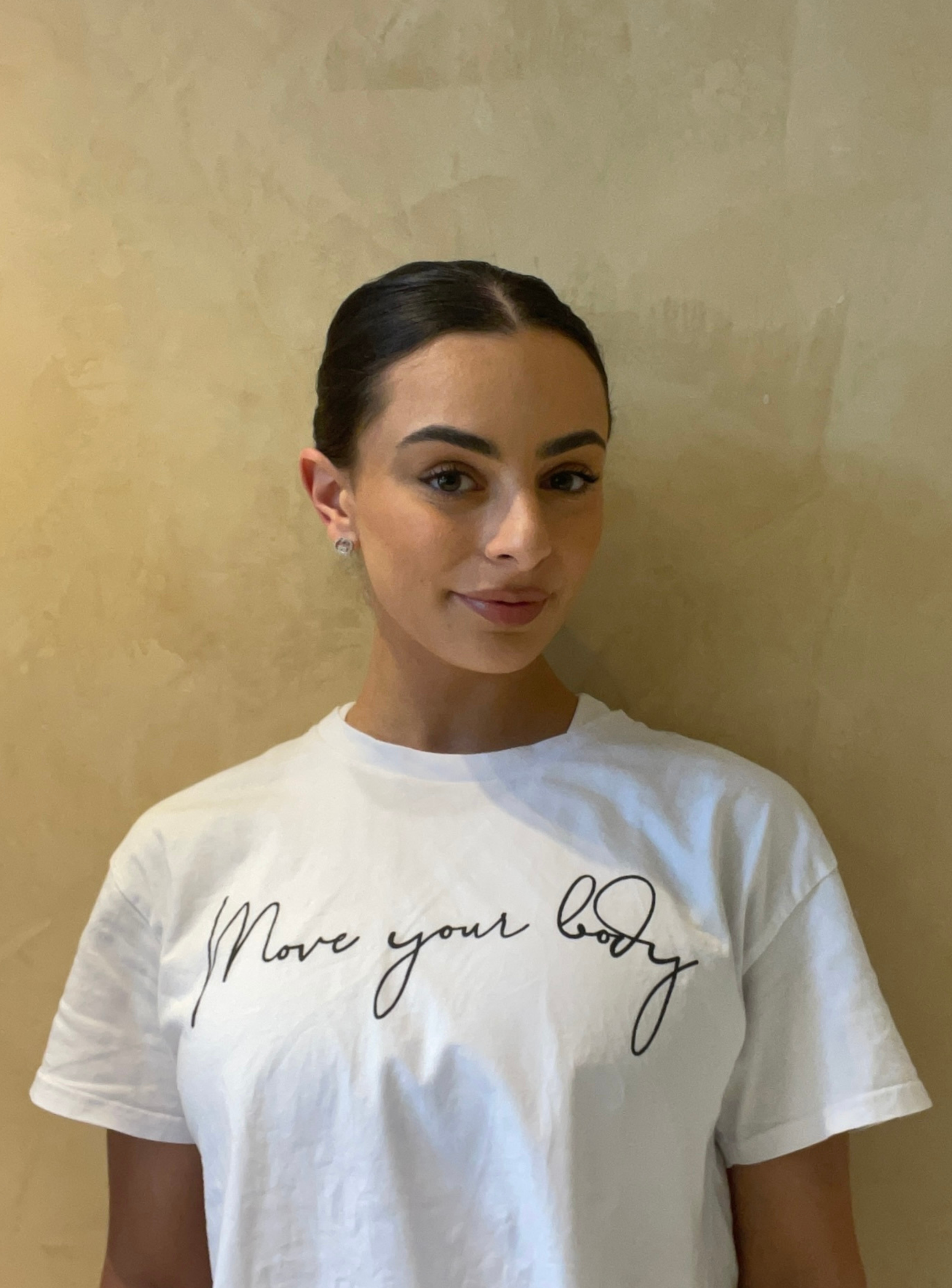 Woman with long hair wearing a white t-shirt, standing against a beige wall.