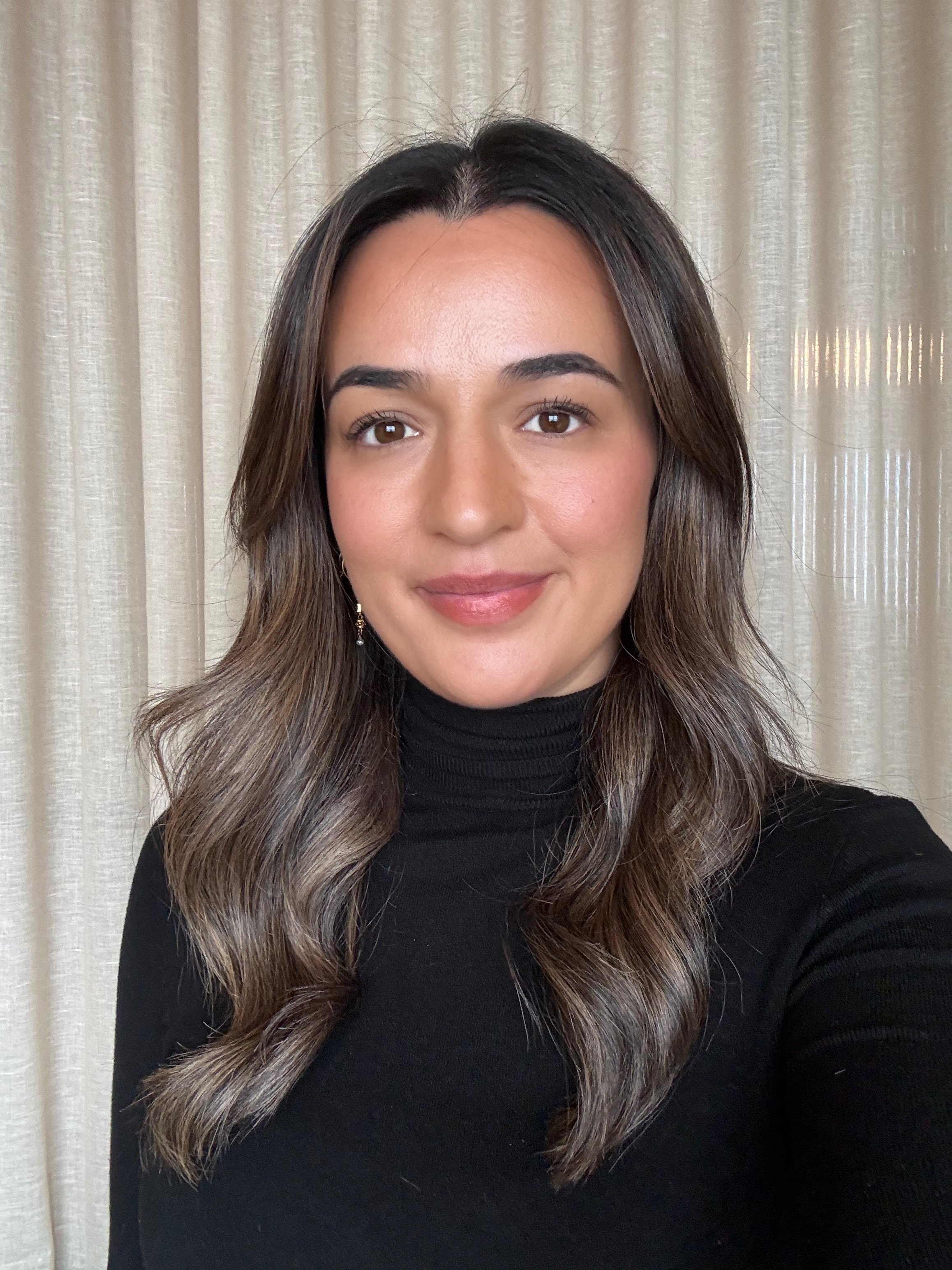 A woman with long brown hair, wearing a leopard print blouse, smiling and standing against a light gray textured wall.