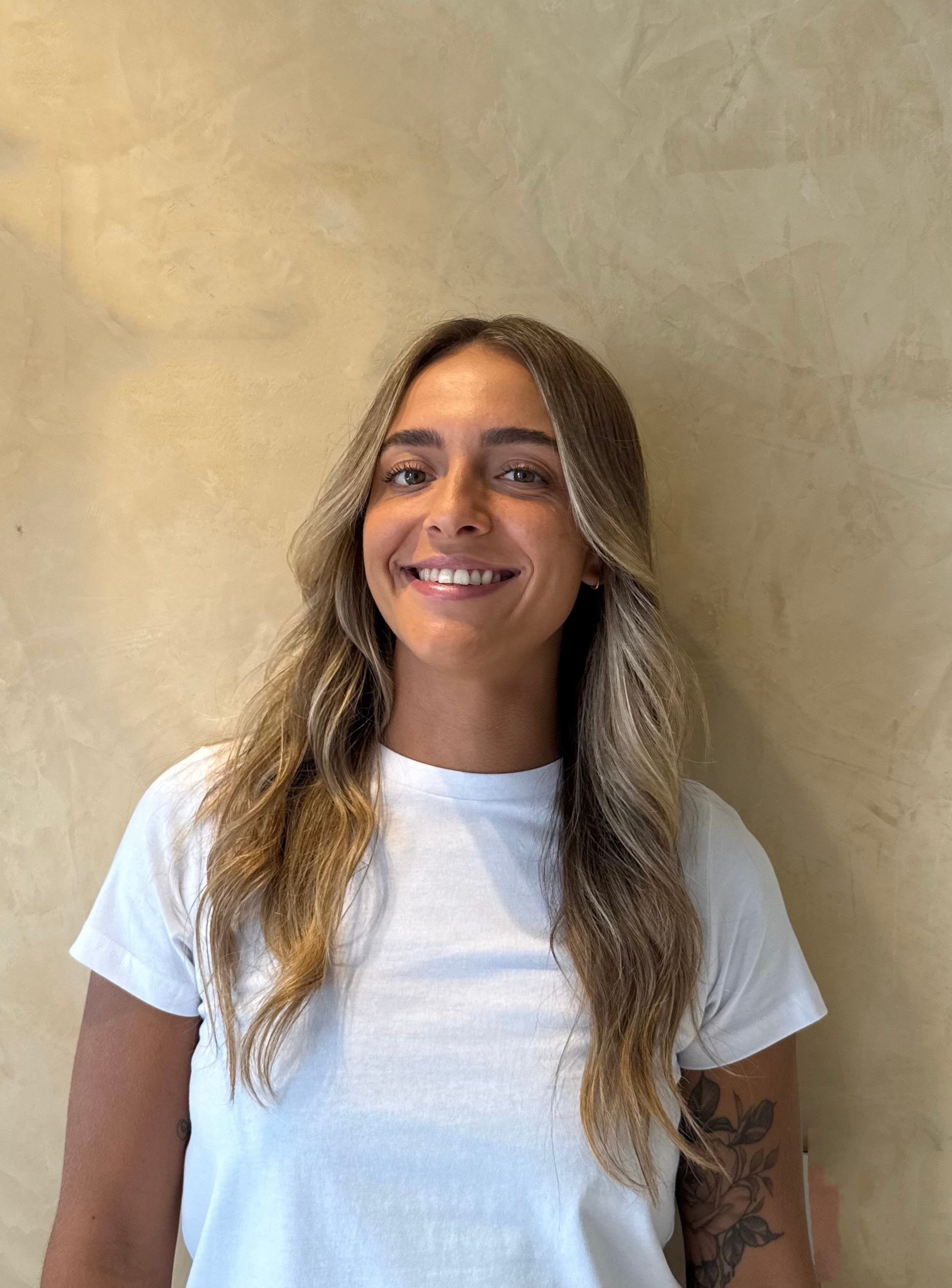 Smiling woman with glasses and long hair wearing a white shirt standing against a beige background.
