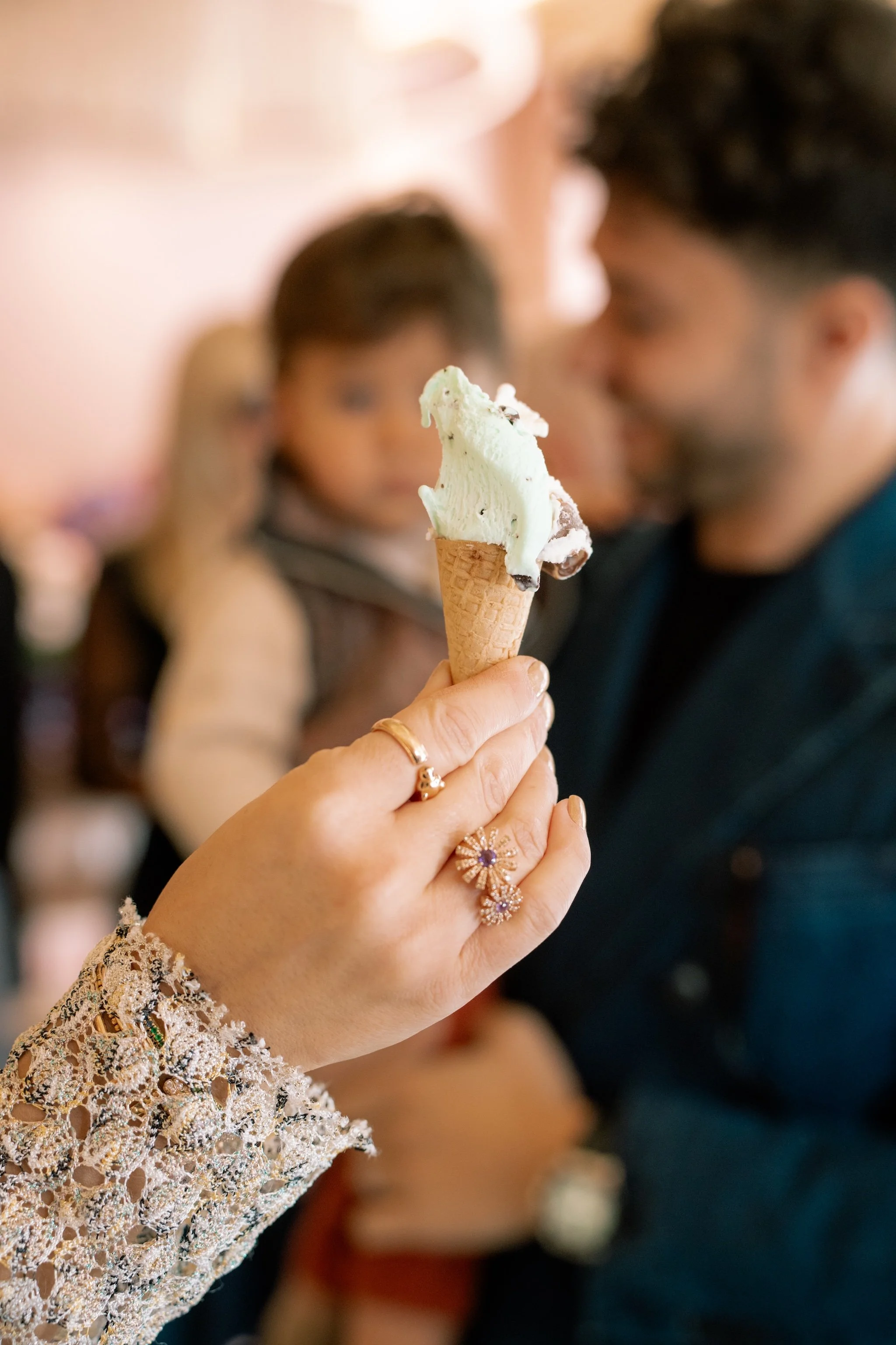 Nahaufnahme einer Hand mit mehreren Ringen, die eine Eistüte mit zwei Kugeln Eis hält, im Hintergrund sind ein Mann und ein Kind in einem warm beleuchteten Raum, vermutlich eine Eisdiele.