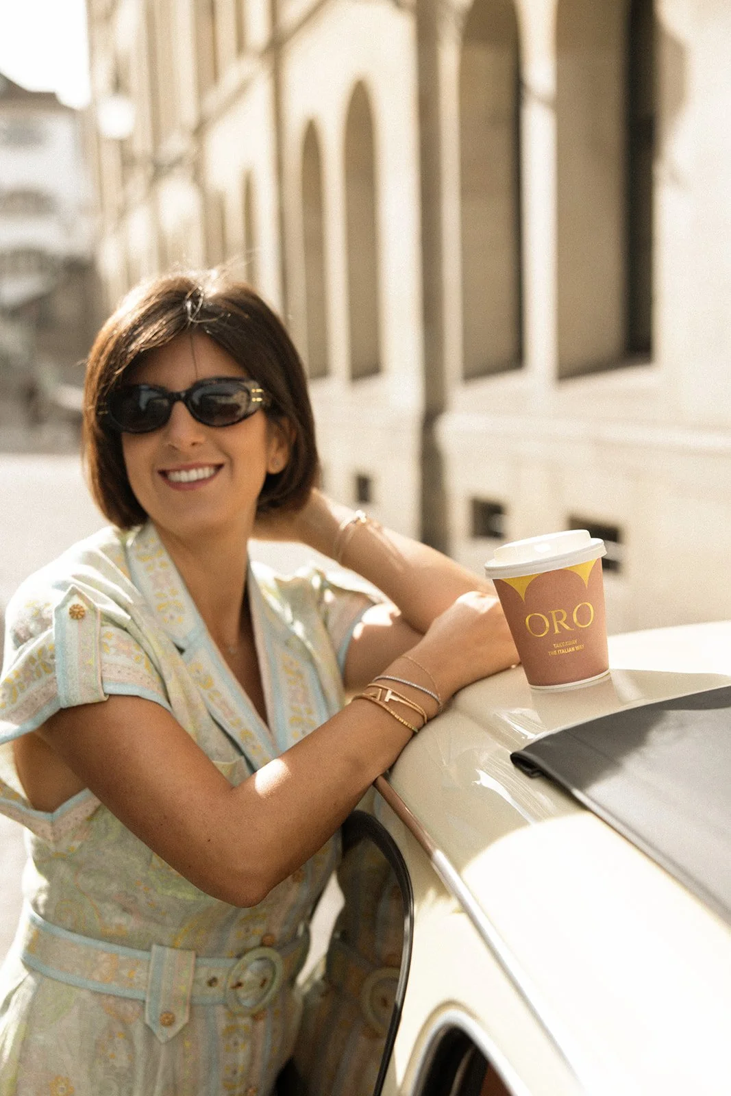 Eine lachende Frau mit dunklen Haaren, Sonnenbrille, armbändern und einem gestreiften Kleid lehnt an einem weißen Auto mit Kaffee to-go-Becher auf dem Dach, in einer sonnenverwöhnten Stadtstraße.