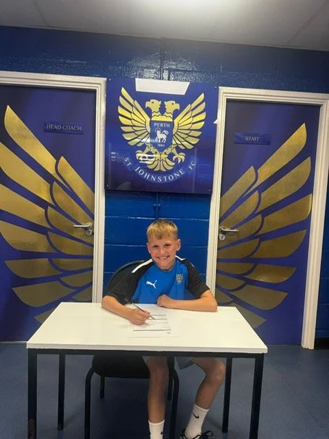 A young boy sitting at a white table, smiling, in front of a blue wall with gold eagle wings and a logo that says 'Perth St. Johnstone FC.' There are two doors labeled 'Head Coach' and 'Start' on either side of the logo.