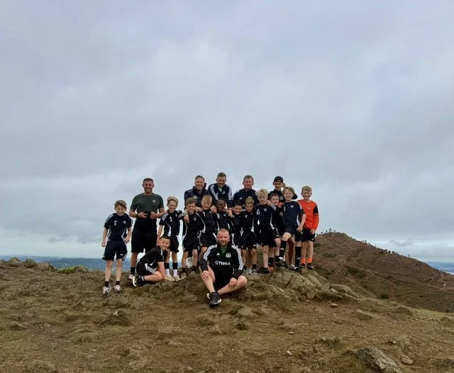 Group of children and adults posing for a photo on a rocky hilltop during overcast weather, some wearing sports gear.