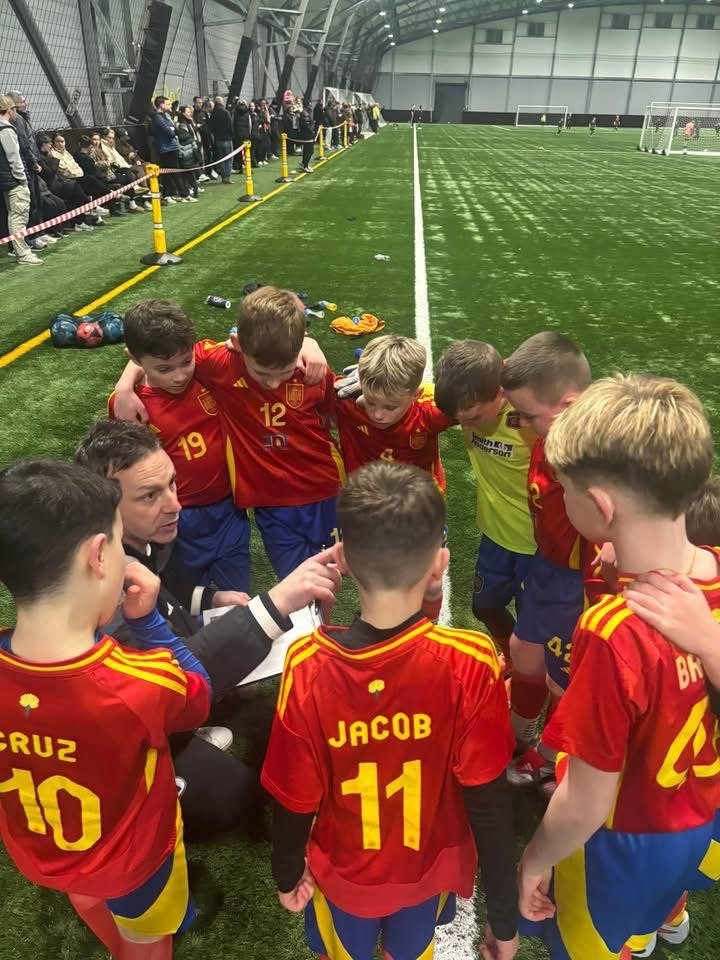A youth soccer team in red jerseys huddles around their coach during a timeout inside an indoor soccer facility, with spectators watching from the sidelines.
