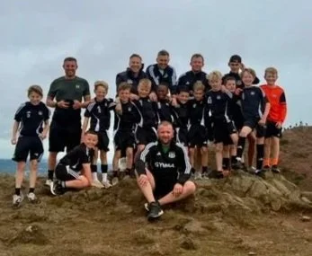 Group of boys and men in black sports uniforms posing outdoors on a hilltop during daytime.