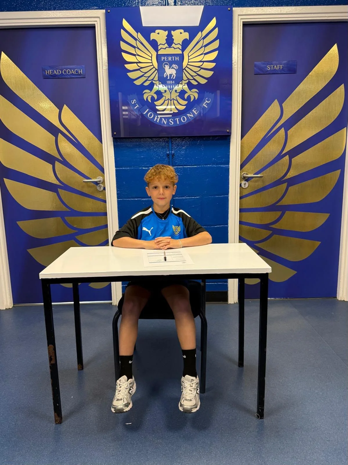 Young boy sitting at a desk in front of a large blue and gold emblem of St. Johnstone Football Club, with doors on either side labeled 'Head Coach' and 'Staff'. The boy is wearing a blue and black sports jersey, black shorts, and white sneakers.