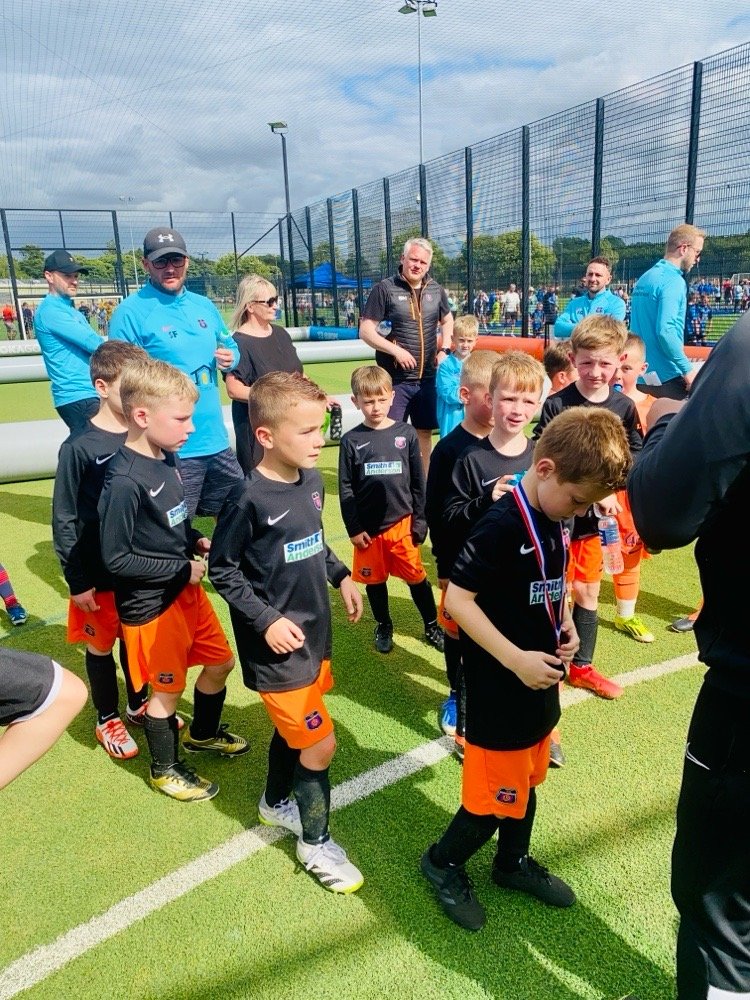 Kids in black soccer jerseys and orange shorts standing on a green soccer field during a sports event. Adults are watching in the background near a fence, with a cloudy sky overhead.