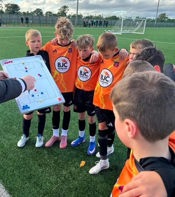 Kids in orange soccer jerseys gathered around a coach who is pointing at a tactics board during a soccer practice on an outdoor field.